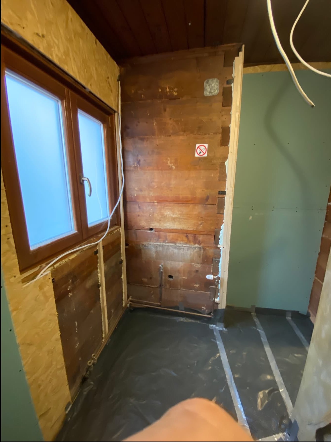 Wnętrze w trakcie remontu: drewniana ściana z instalacjami, okno z widoczną konstrukcją, fragment płyty karton-gips i czarna folia na podłodze. Widoczne etapy prac wykończeniowych.