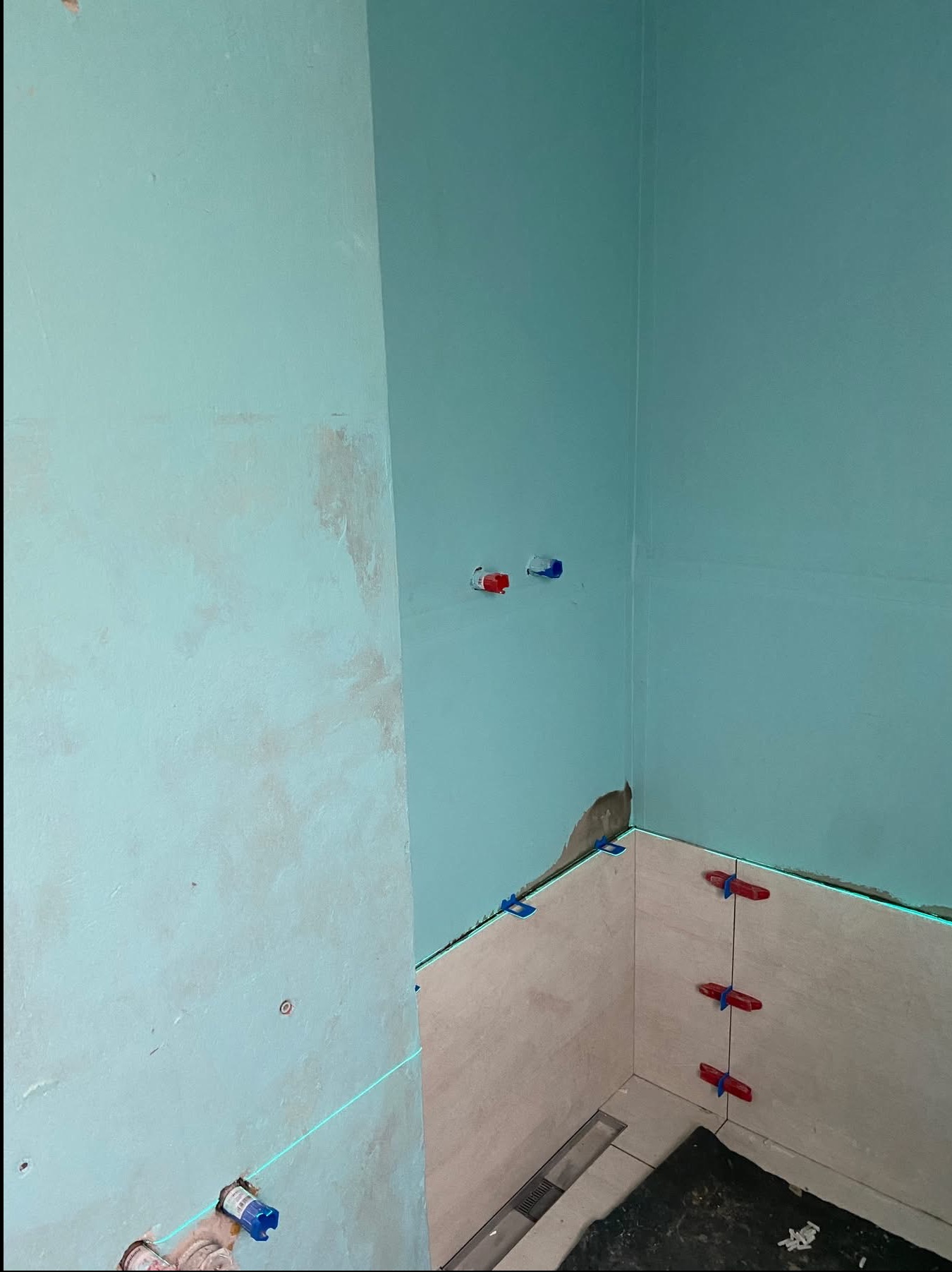 Łazienka w trakcie remontu: częściowo wyłożone płytki, widoczne krzyżyki dystansowe i poziomica laserowa. Ściany w kolorze turkusowym, przygotowane do dalszych prac.