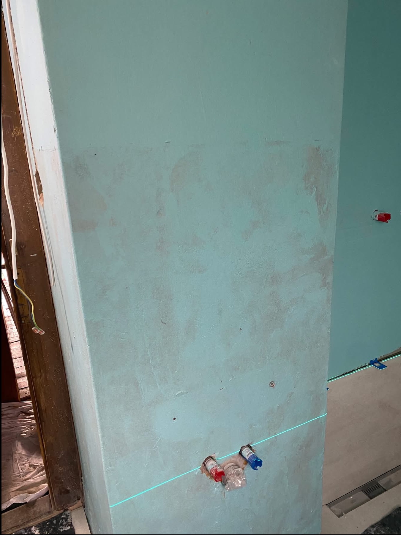 Nierówna, pomalowana na błękitno ściana z widocznymi śladami po instalacji wodnej, wyznaczona poziomą linią lasera. Fragment podłogi z odpływem liniowym.