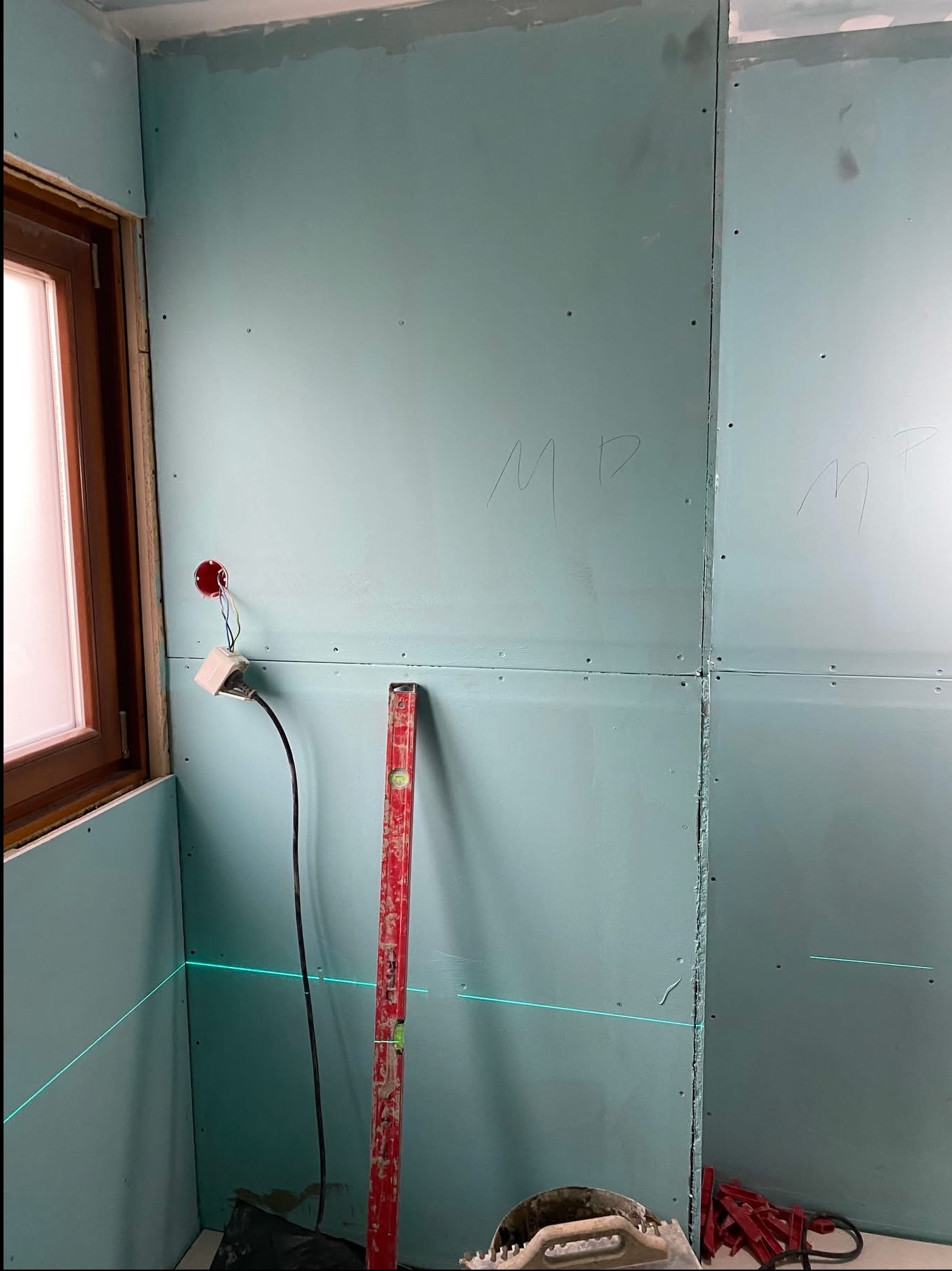 Montaż płyt karton-gips na ścianie z widocznym poziomowaniem laserowym, poziomica i wystającym kablem elektrycznym. Ściany przygotowane do dalszych prac wykończeniowych.