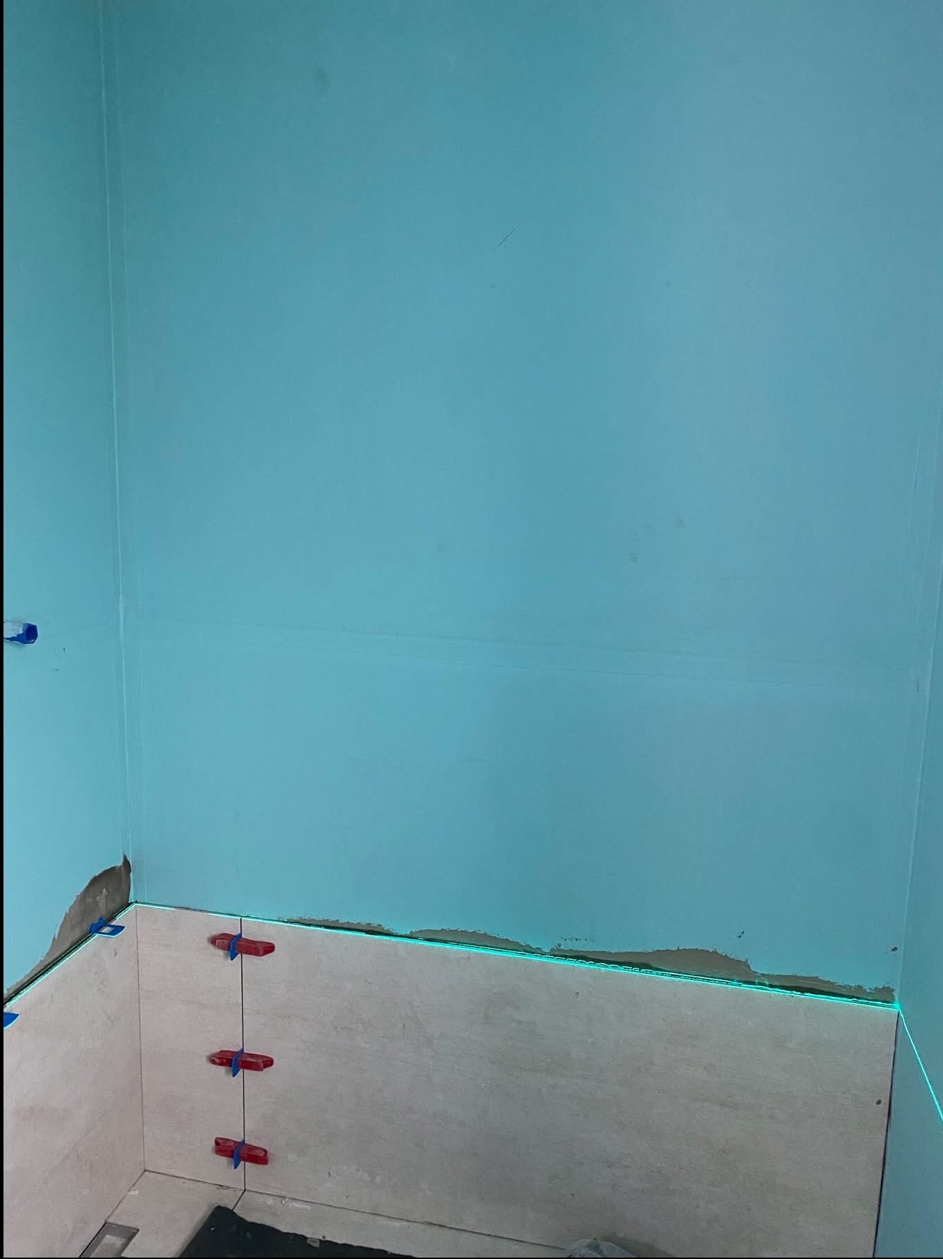 Układanie płytek w rogu pomieszczenia z użyciem poziomicy laserowej. Widoczne krzyżyki dystansowe i fragment ściany pomalowanej na turkusowo. Prace glazurnicze w toku.