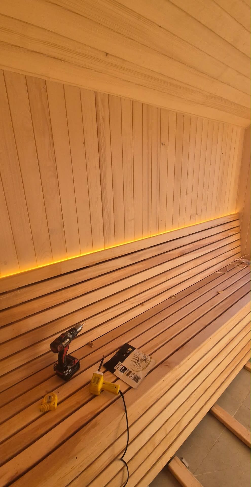 Drewniana sauna w trakcie budowy z podświetleniem LED, widoczne narzędzia: wkrętarka, lutownica, miarka i opakowanie kabli. Ściany i ławki z jasnego drewna.