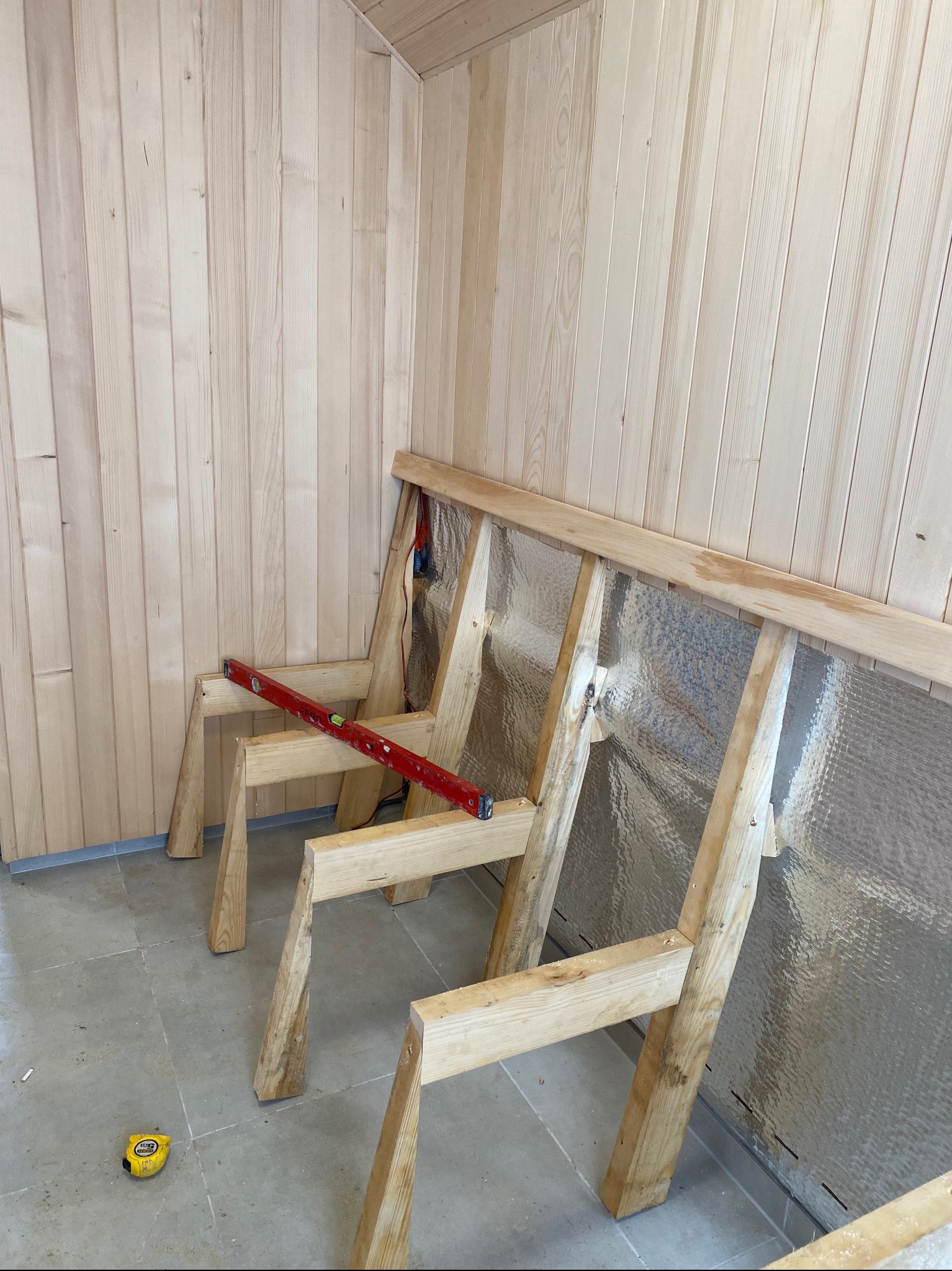 Drewniana konstrukcja szkieletowa z izolacją z folii aluminiowej przy ścianie z boazerii. Poziomica i miarka leżą na podłodze. Prace wykończeniowe wewnątrz pomieszczenia.