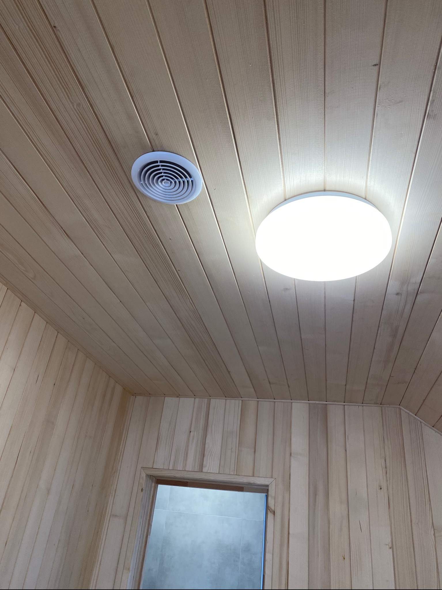 Drewniana boazeria na suficie i ścianach, okrągła lampa sufitowa i kratka wentylacyjna. Widoczny fragment drzwi. Jasne, naturalne drewno.