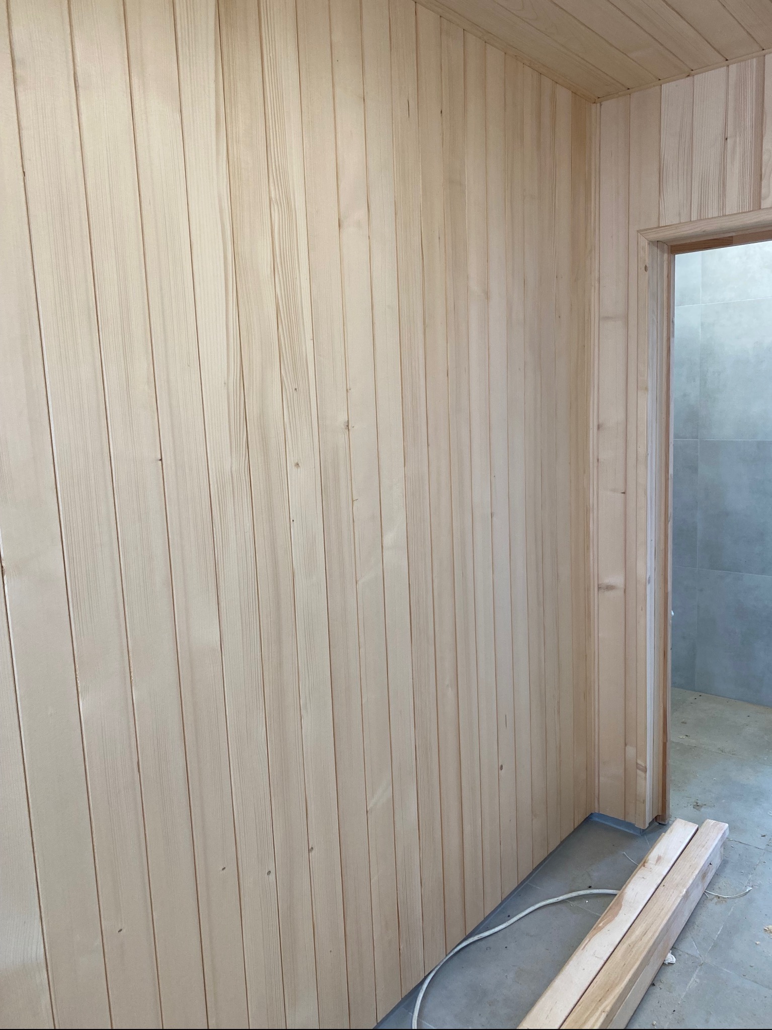 Jasne, drewniane panele boazeryjne pokrywają ściany i sufit, tworząc przytulne wnętrze. Widoczne fragmenty podłogi i drzwi do innego pomieszczenia z szarymi płytkami.