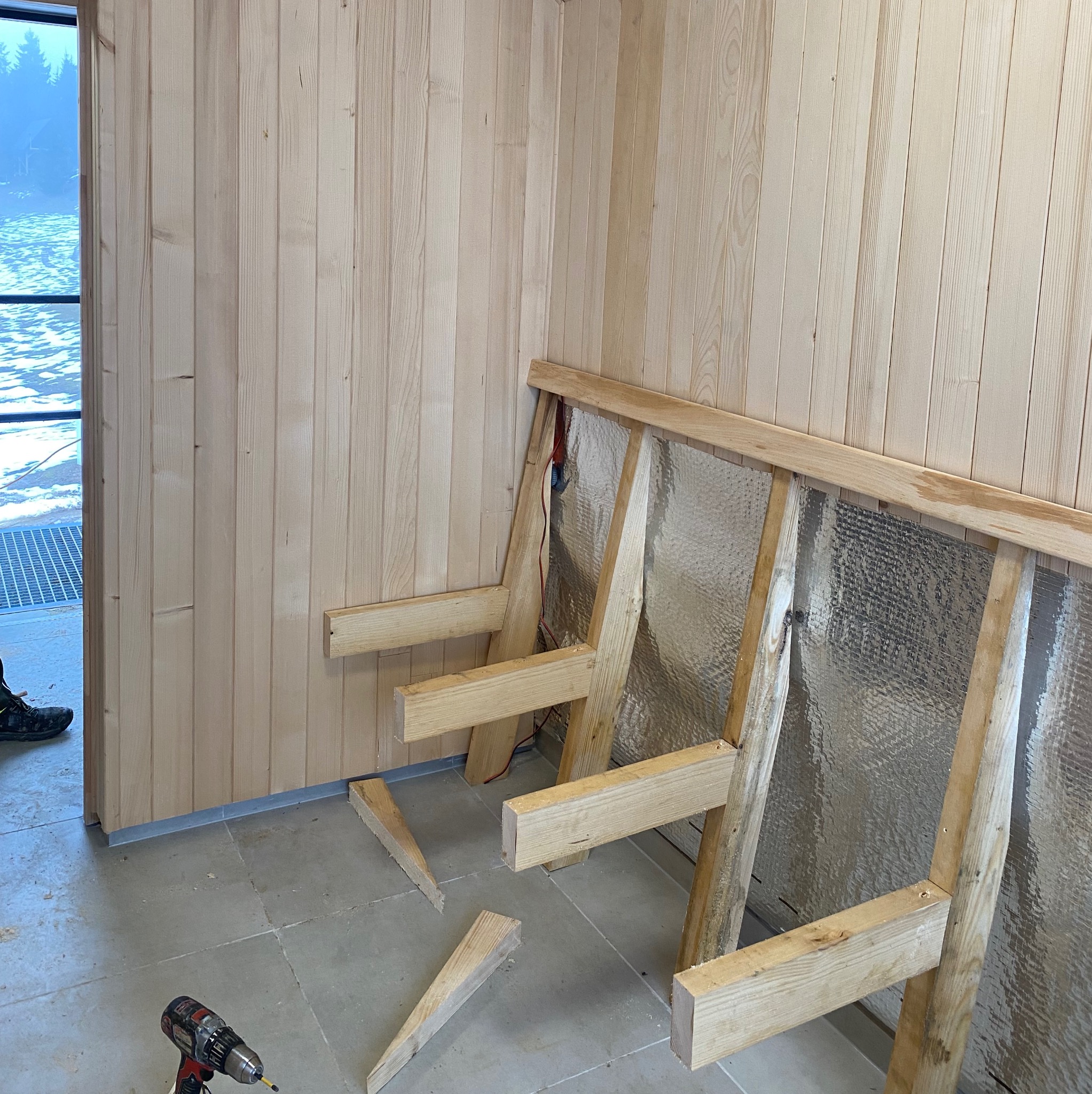 Montaż boazerii drewnianej na ścianie z izolacją, widoczny szkielet konstrukcji i wkrętarka. Fragment widoku na zewnątrz przez okno. Prace wykończeniowe wewnątrz budynku.