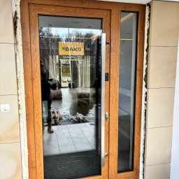 STOLARKA OKIENNO-DRZWIOWA -DAMIAN DRUSZCZ - Drewniane drzwi wejściowe z szybą, zamontowane w jasnej elewacji. Widoczne pianowanie uszczelniające. Na szybie naklejka producenta DAKO.