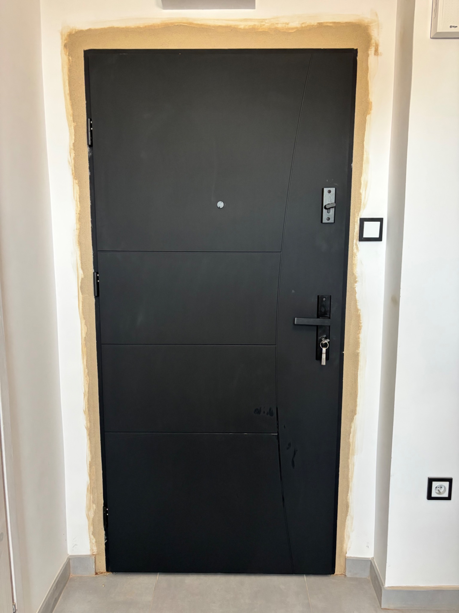 Nowoczesne, czarne drzwi wejściowe z ościeżnicą w trakcie wykończenia, widoczne klucze w zamku, na tle białej ściany z gniazdkami elektrycznymi.