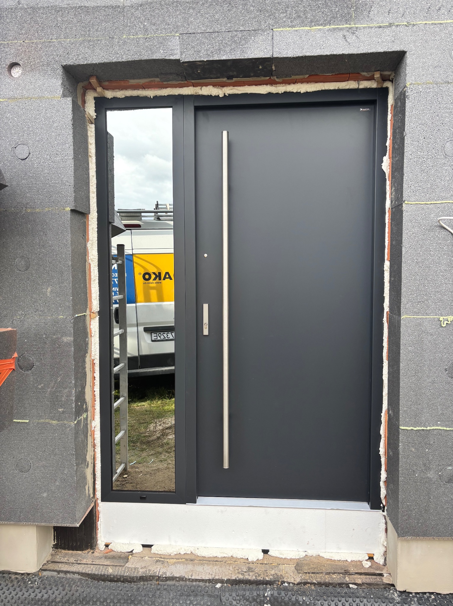 Zamknięte, grafitowe drzwi wejściowe z długim, srebrnym uchwytem, zamontowane w surowym otworze z szarych bloczków i pianki montażowej. Widoczne odbicie w szybie.