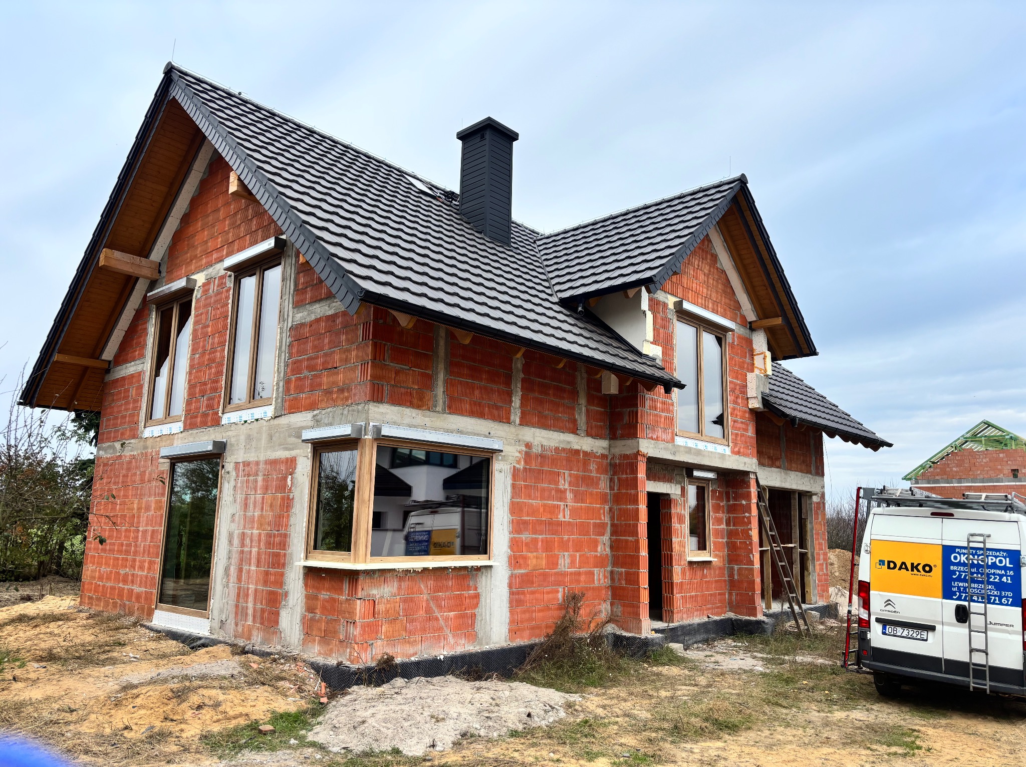 Dom w trakcie budowy z zamontowanymi oknami, widoczny z zewnątrz. Na posesji stoi samochód dostawczy z reklamą firmy okiennej.