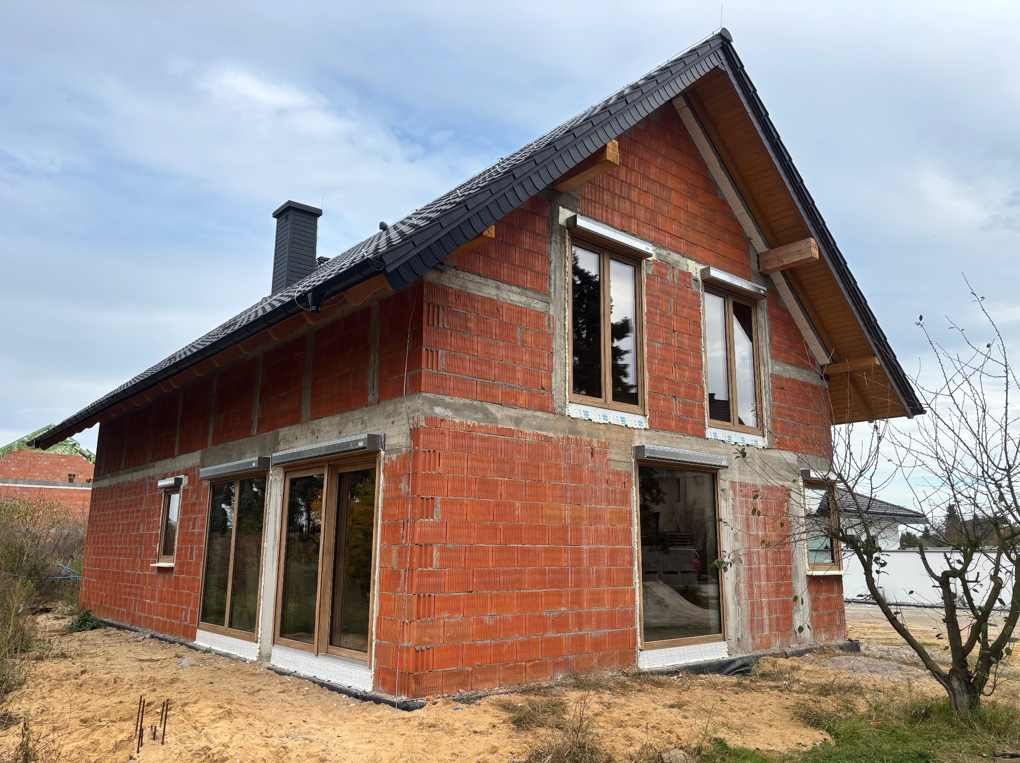 Surowy budynek z czerwonej cegły z zamontowanymi oknami i roletami zewnętrznymi, czarnym dachem i kominem, w trakcie budowy, na tle pochmurnego nieba.