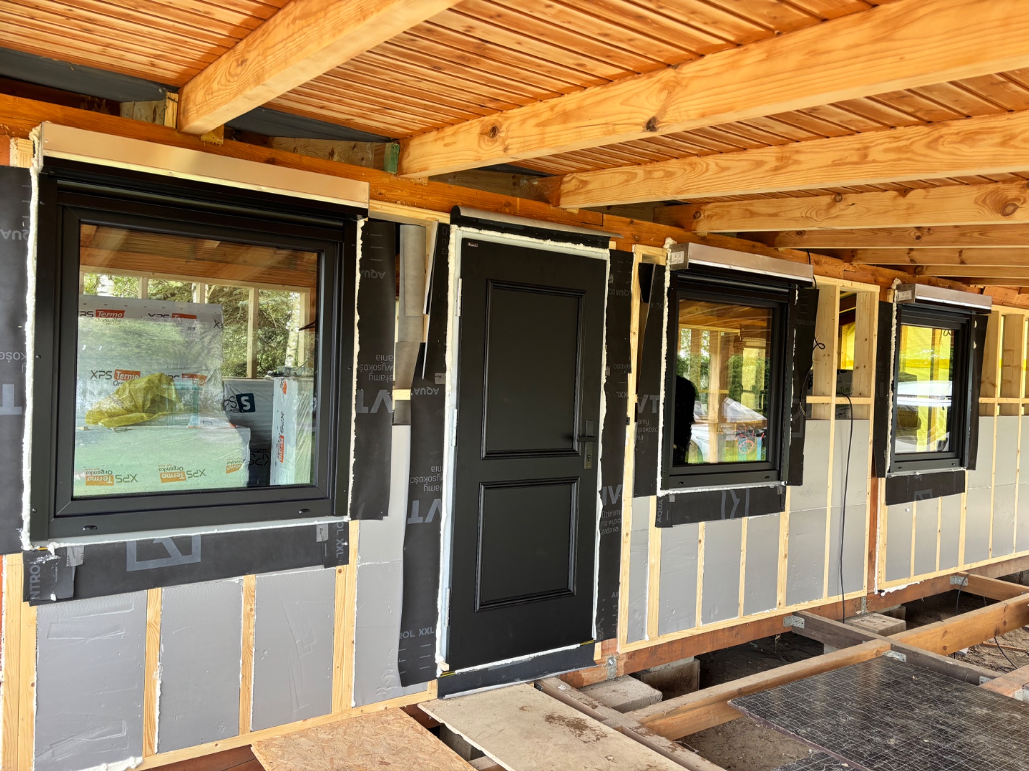 Częściowo ocieplona ściana z zamontowanymi czarnymi oknami i drzwiami. Widoczne drewniane elementy konstrukcyjne i materiały izolacyjne. Prace wykończeniowe w trakcie.