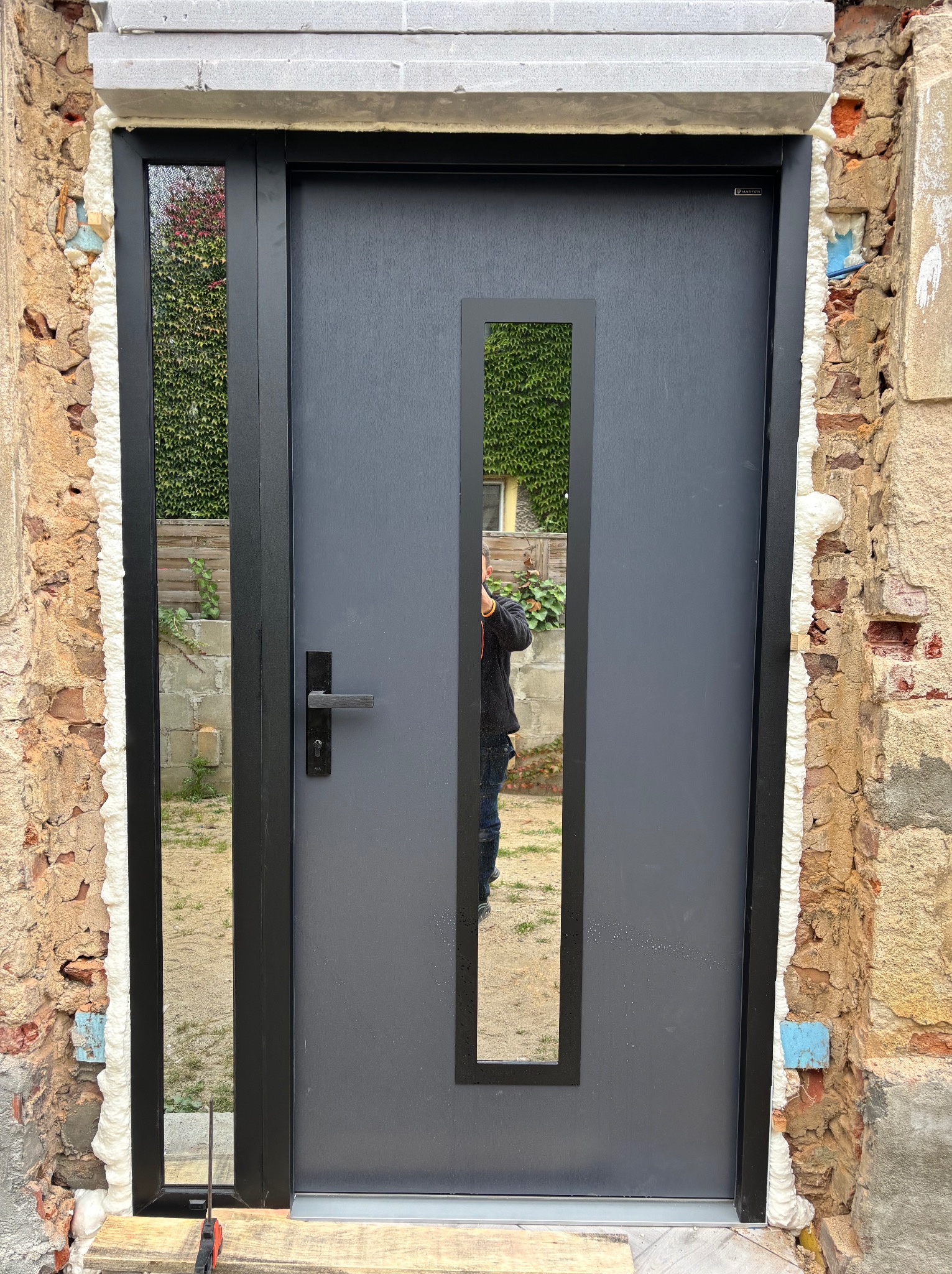 Szare drzwi wejściowe z długim, wąskim przeszkleniem, osadzone w surowym murze z pianką montażową. Widoczne narzędzia i odbicie osoby w szybie.