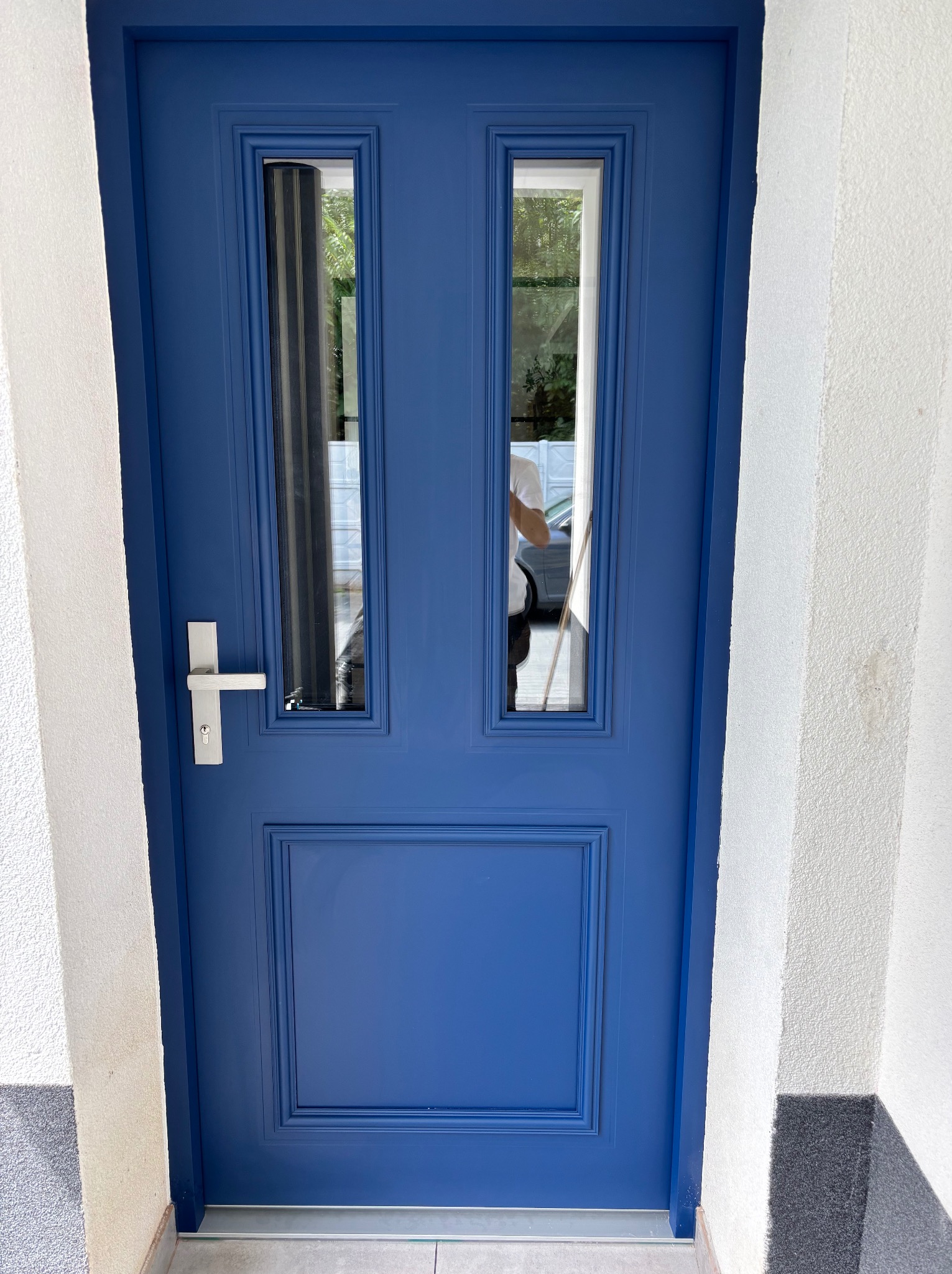 Niebieskie drzwi wejściowe z dwoma szybami i srebrną klamką, zamontowane w białej ścianie. Widoczne odbicie osoby w szybie.