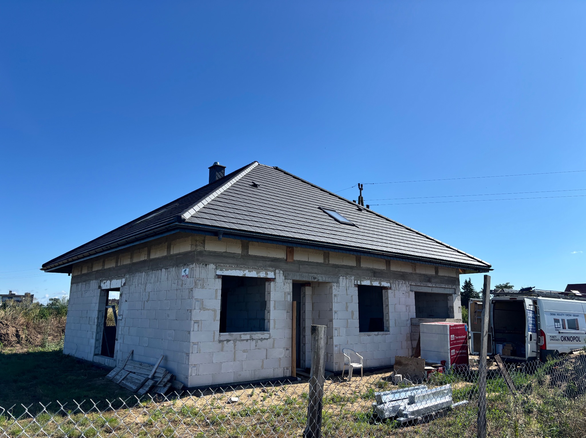 Surowy stan domu jednorodzinnego z pustaków, z widocznym dachem z dachówki ceramicznej i otworami okiennymi, na tle błękitnego nieba i częściowo widocznego samochodu dostawczego.