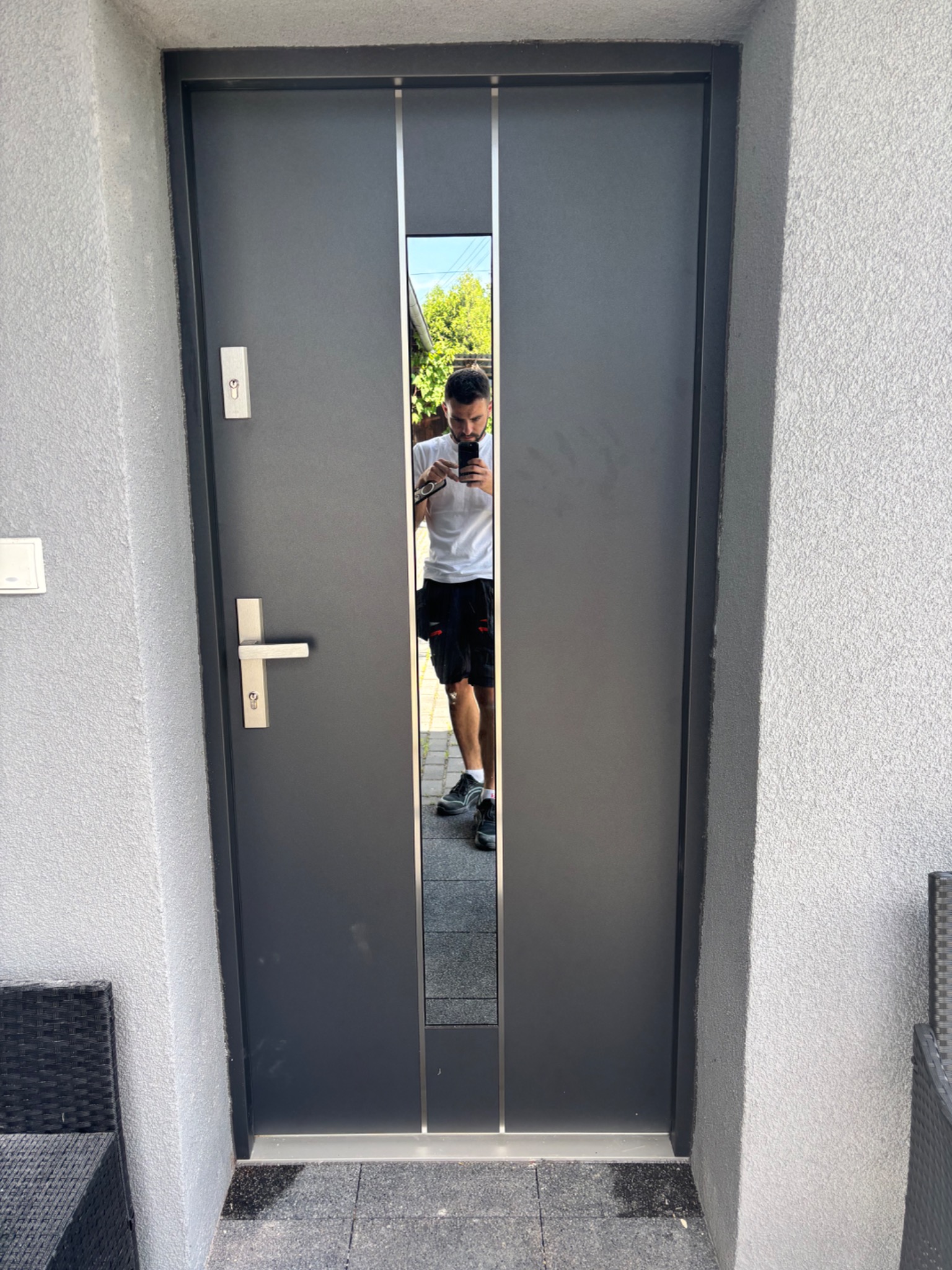 Nowoczesne, grafitowe drzwi wejściowe z lustrzanym panelem, zamontowane w jasnej elewacji. Widoczne detale: klamka, zamek, fragment odbicia osoby robiącej zdjęcie.