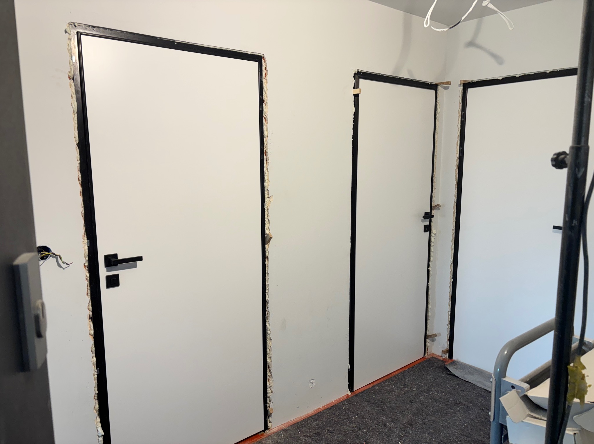 Montaż trzech białych drzwi z czarną ościeżnicą w surowym stanie. Widoczne szczeliny wypełnione pianką montażową i wystające kable elektryczne.
