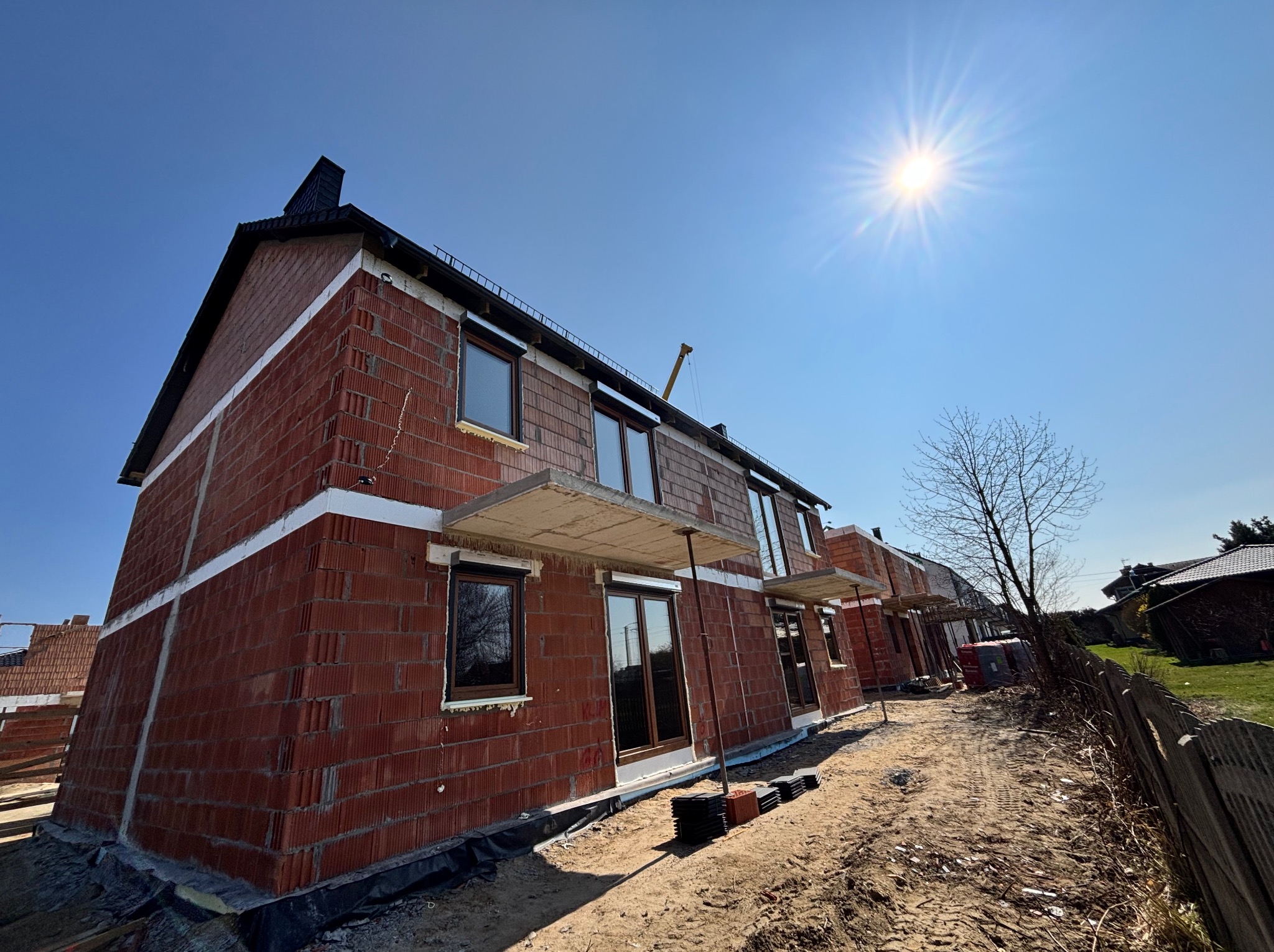 Budowa domu z czerwonej cegły, widoczne okna z brązowymi ramami i przygotowane otwory na drzwi. Słoneczny dzień, niebieskie niebo. Stan surowy otwarty.