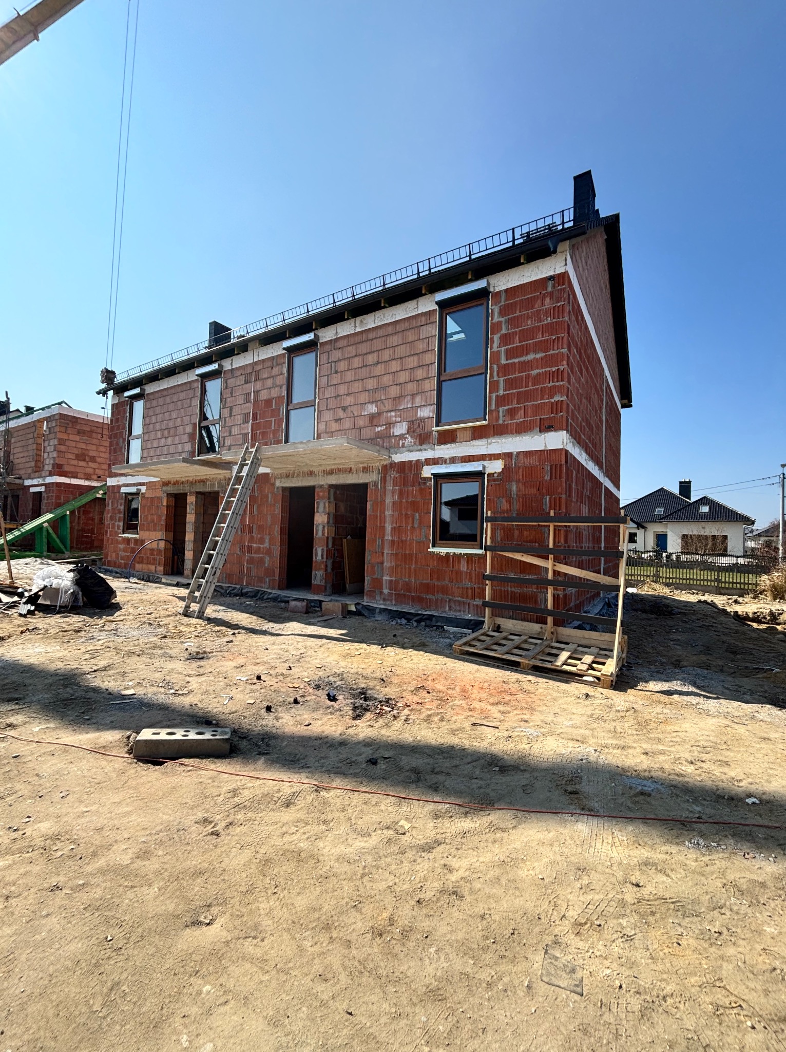 Surowy stan bliźniaka z czerwonej cegły, widoczne okna i drabina oparta o ścianę. Budowa w toku, ziemia wokół, błękitne niebo w tle.