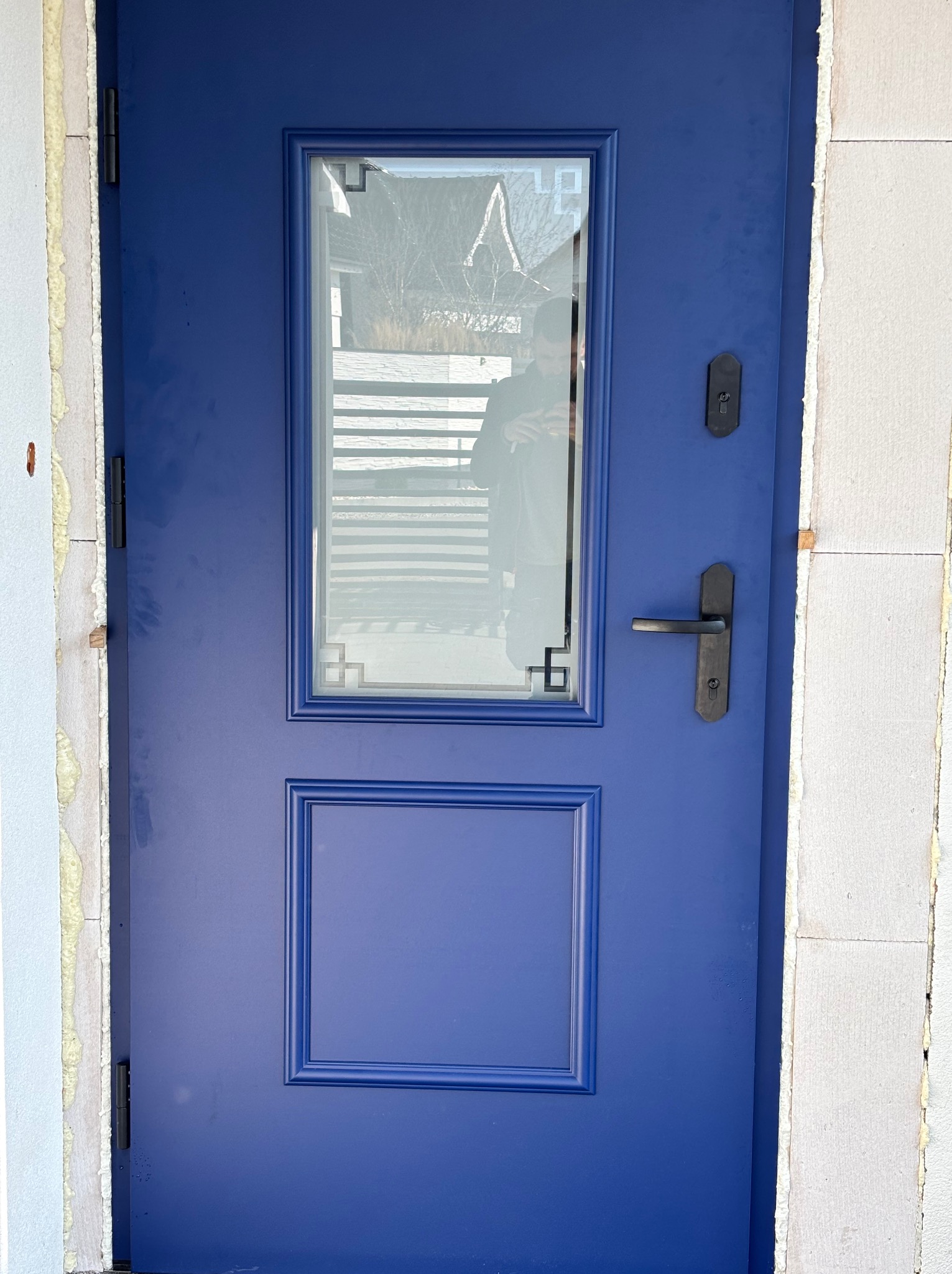 Niebieskie drzwi wejściowe z szybą i ozdobnymi ramkami, zamontowane w surowym otworze, widoczna pianka montażowa i bloczki. Nowoczesny design.