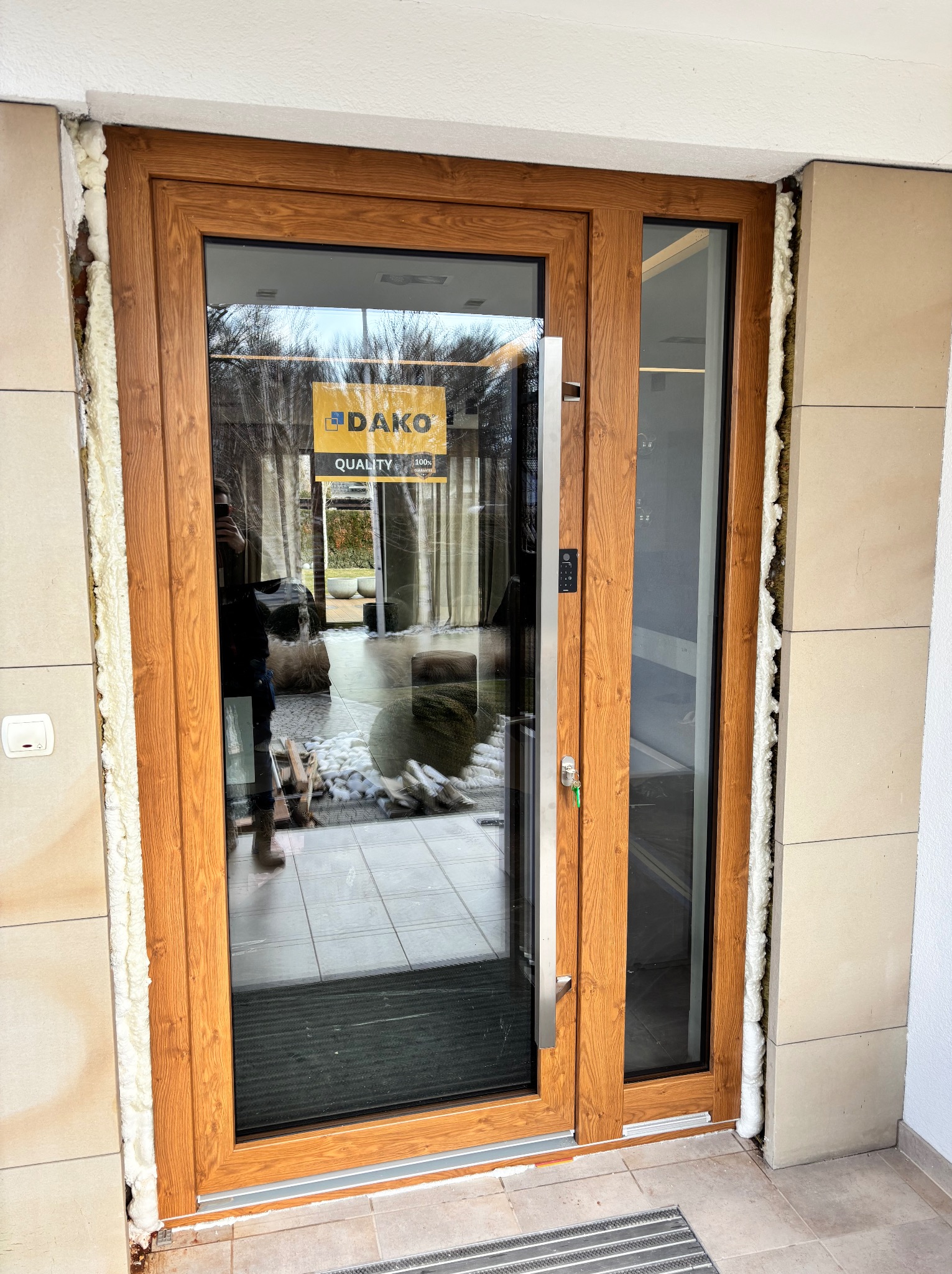 Drewniane drzwi wejściowe z szybą, zamontowane w jasnej elewacji. Widoczne pianowanie uszczelniające. Na szybie naklejka producenta DAKO.