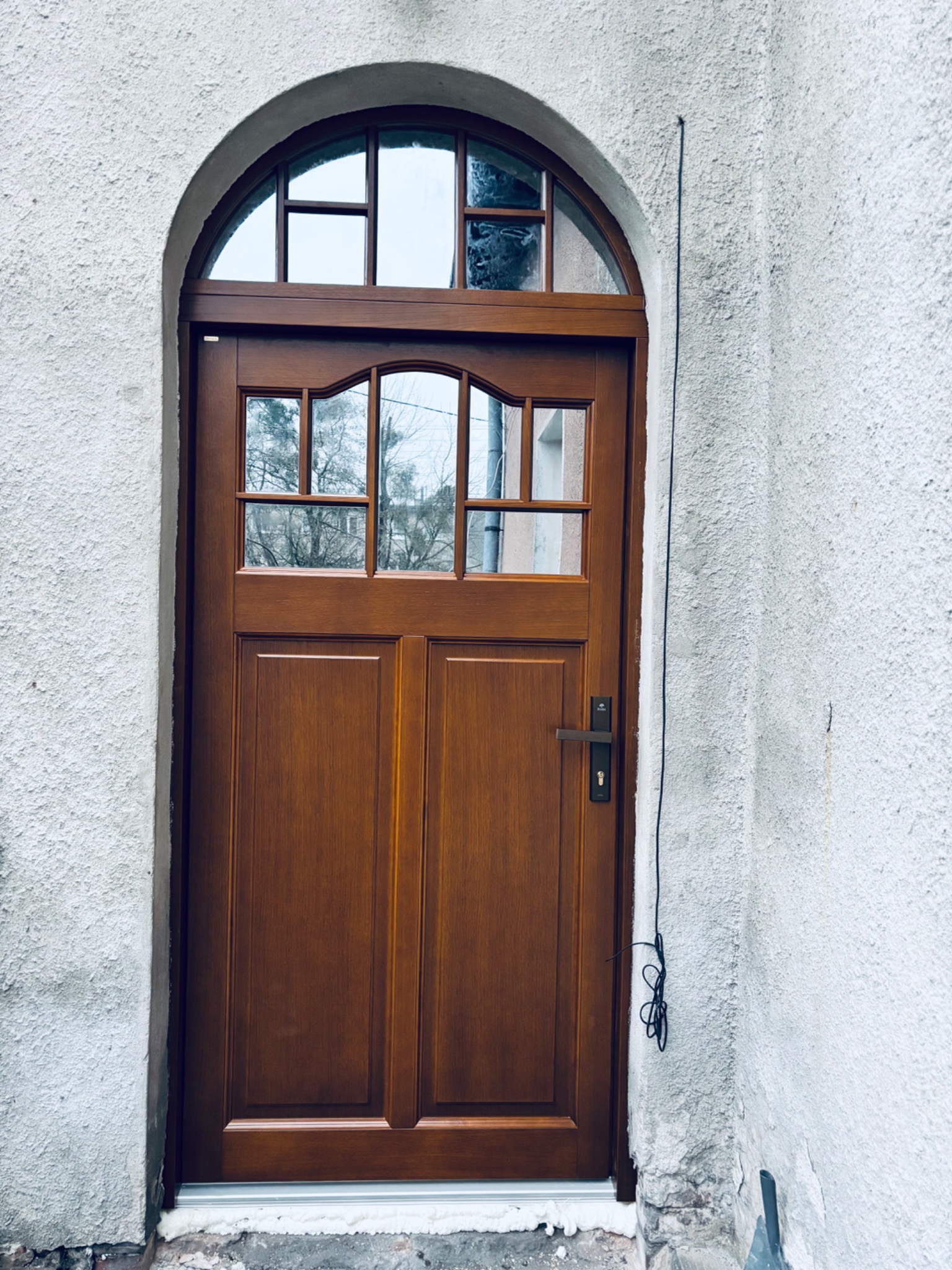 Drewniane drzwi wejściowe z łukowym przeszkleniem, zamontowane w Brzegu. Widoczna faktura ściany i uszczelnienie wokół ościeżnicy.