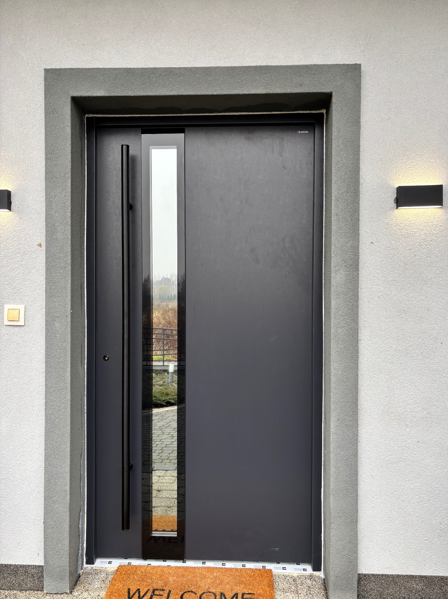 Nowoczesne, grafitowe drzwi wejściowe z długim uchwytem i pionowym przeszkleniem, zamontowane w szarej elewacji z oświetleniem zewnętrznym.