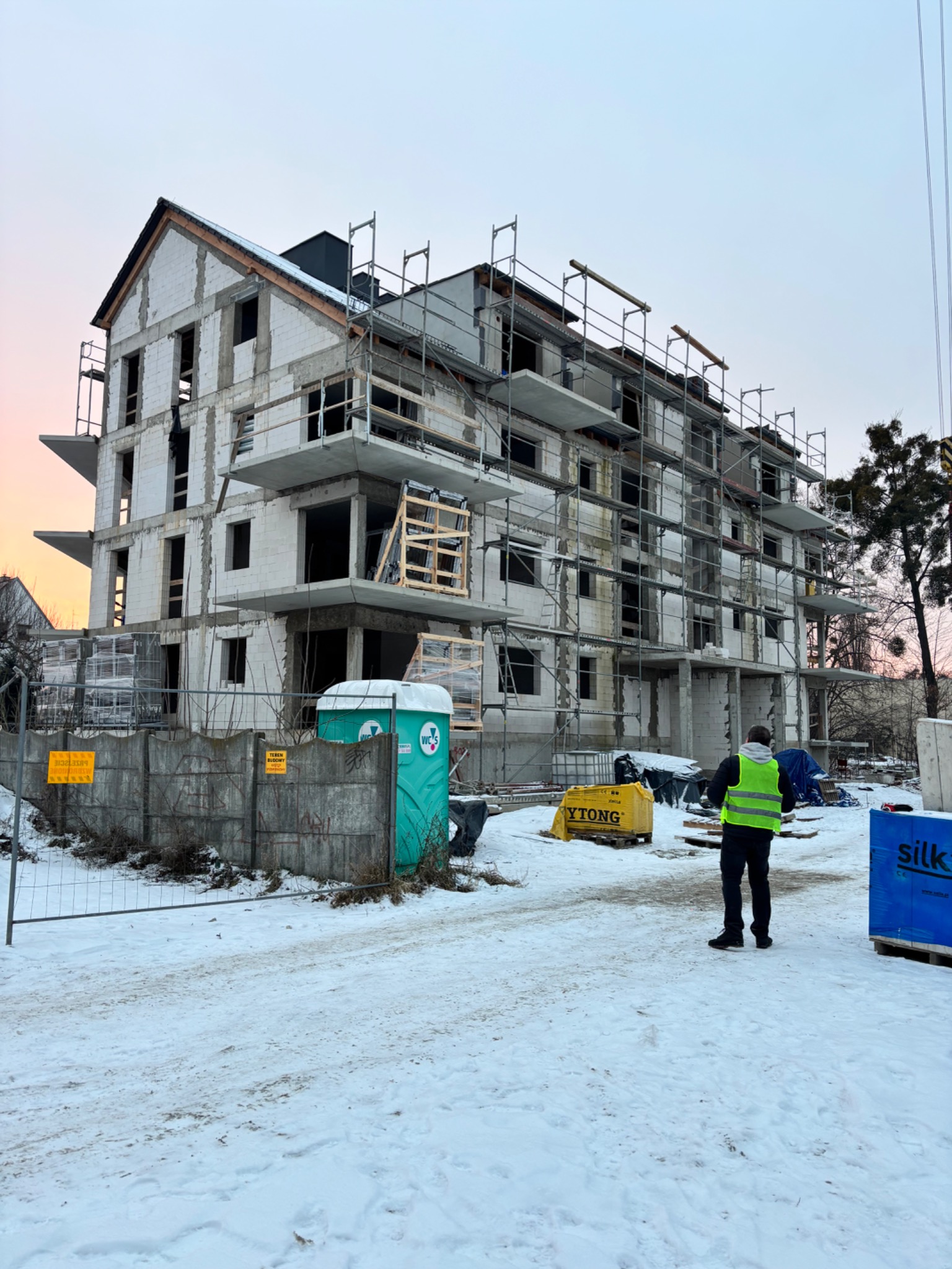 Wznoszony budynek wielorodzinny w zimowej scenerii, z widocznymi rusztowaniami i pracownikiem w odblaskowej kamizelce. Stan surowy, okna i drzwi niezamontowane.