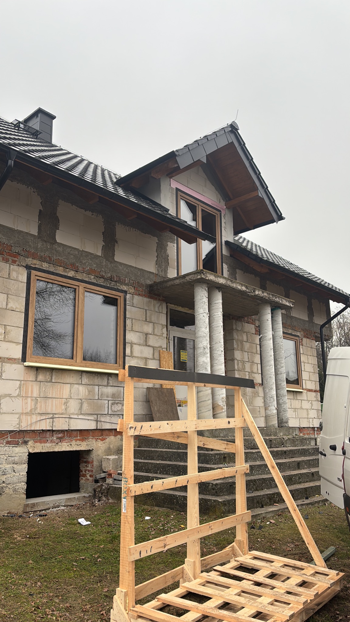 Dom w trakcie budowy z zamontowanymi oknami drewnianymi, widoczne rusztowania i elementy konstrukcyjne. Surowy stan budynku.