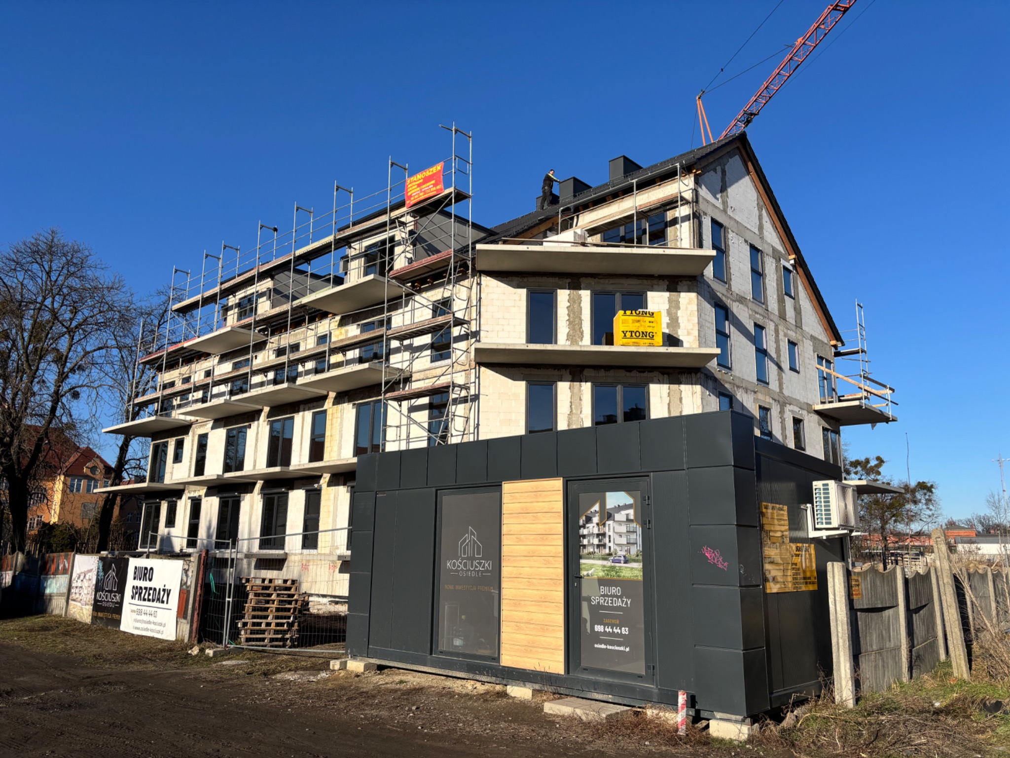 Budowa bloku mieszkalnego w trakcie montażu okien, widoczne rusztowania i biuro sprzedaży z reklamami. Błękitne niebo w tle.
