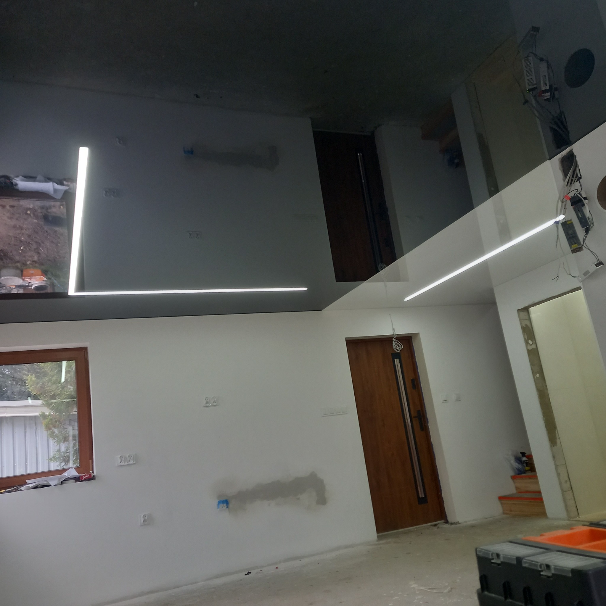 Błyszczący, grafitowy sufit z liniowym oświetleniem LED w nowym wnętrzu. Widoczne okno, drzwi i przygotowane ściany do malowania. Elementy instalacji elektrycznej.