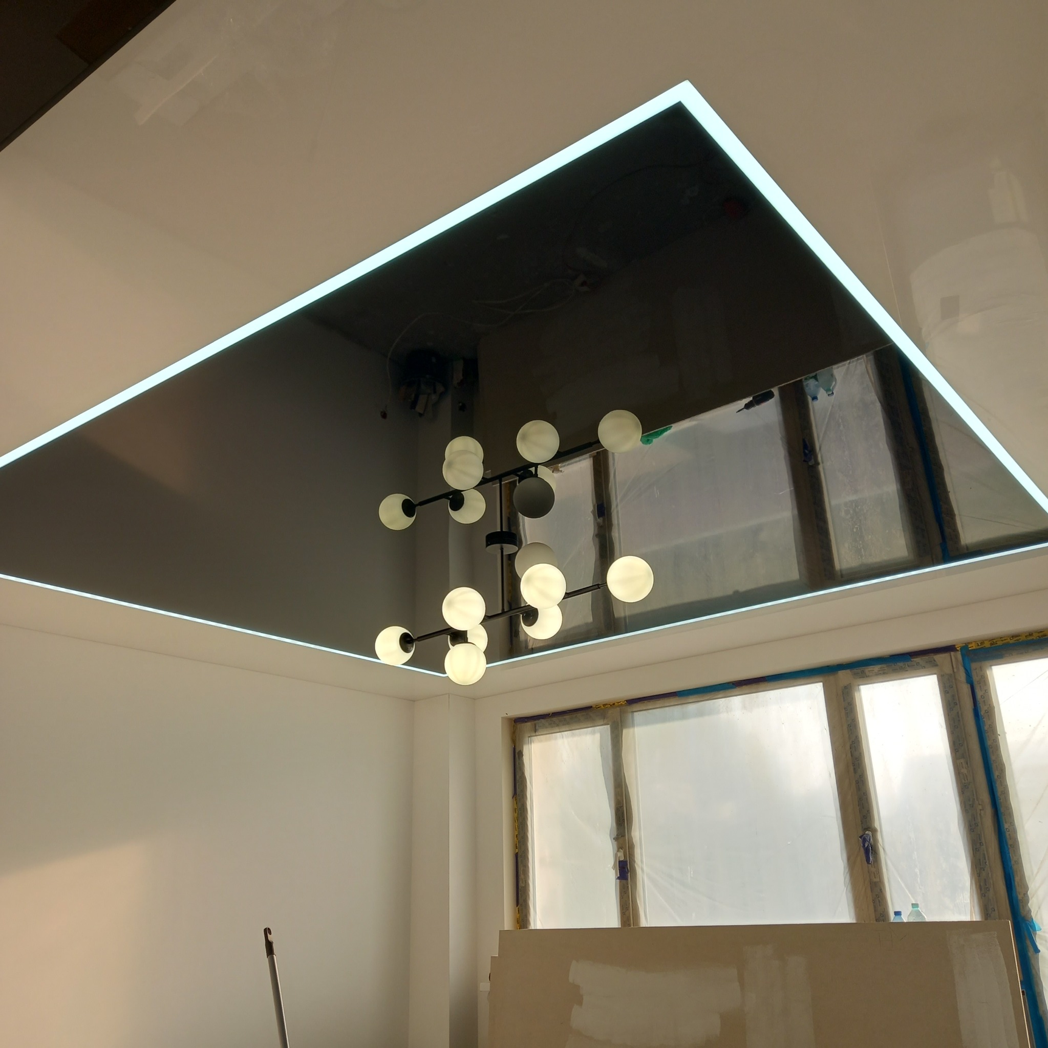 Błyszczący, ciemny sufit z podświetleniem LED w kształcie prostokąta, odbijający nowoczesną lampę kulistą. Widoczne okna zabezpieczone folią malarską.
