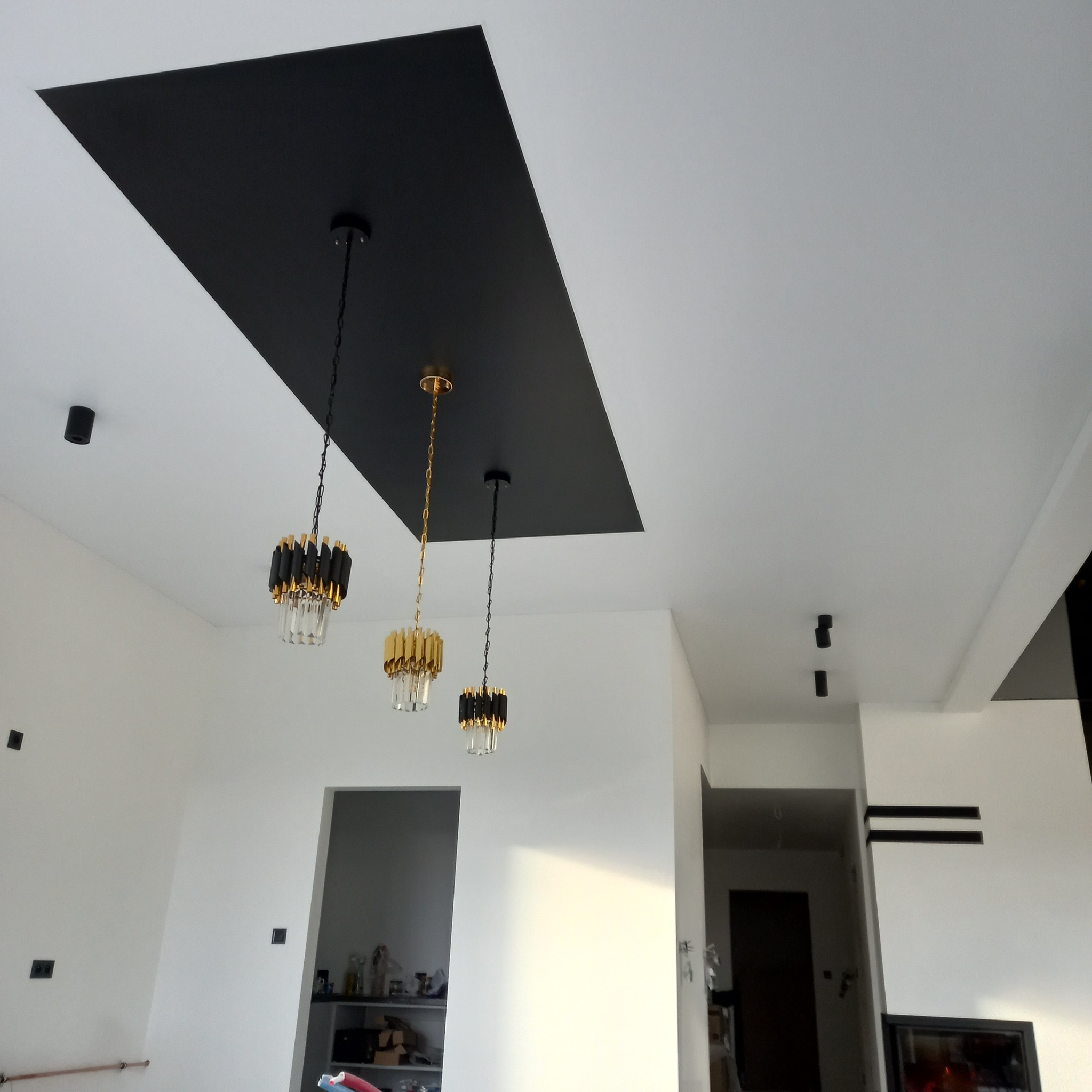 Biały sufit z czarnym, prostokątnym panelem i trzema wiszącymi lampami w złotych i czarnych barwach. Nowoczesne oświetlenie w minimalistycznym wnętrzu.