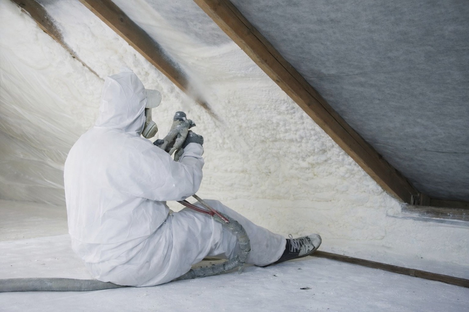 Pracownik w kombinezonie ochronnym aplikuje piankę izolacyjną na poddaszu, widoczne drewniane elementy konstrukcyjne dachu i świeżo nałożona, jasna pianka.