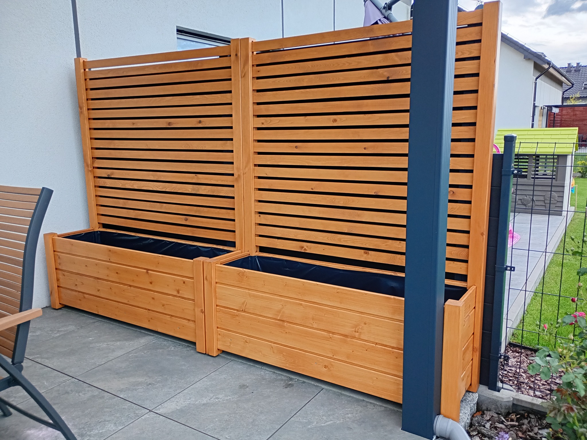 Drewniane donice z przesłonami na tarasie, wyłożonym szarą kostką. W tle widoczny fragment domu i plac zabaw. Ujęcie z boku, z widocznym fragmentem krzesła.