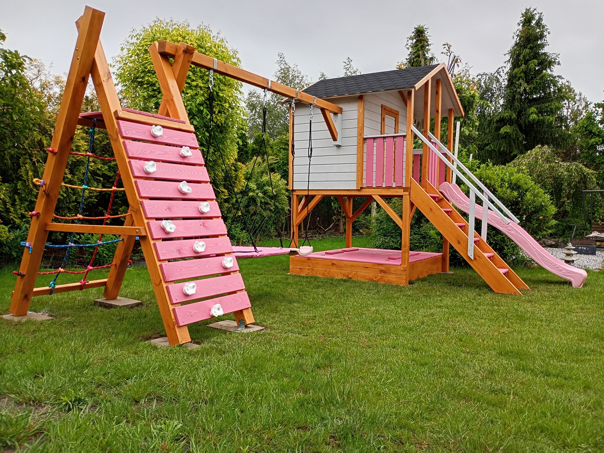 Kolorowy plac zabaw z domkiem, zjeżdżalnią, ścianką wspinaczkową i huśtawką na tle zielonego ogrodu. Drewniana konstrukcja pomalowana na różowo i biało.