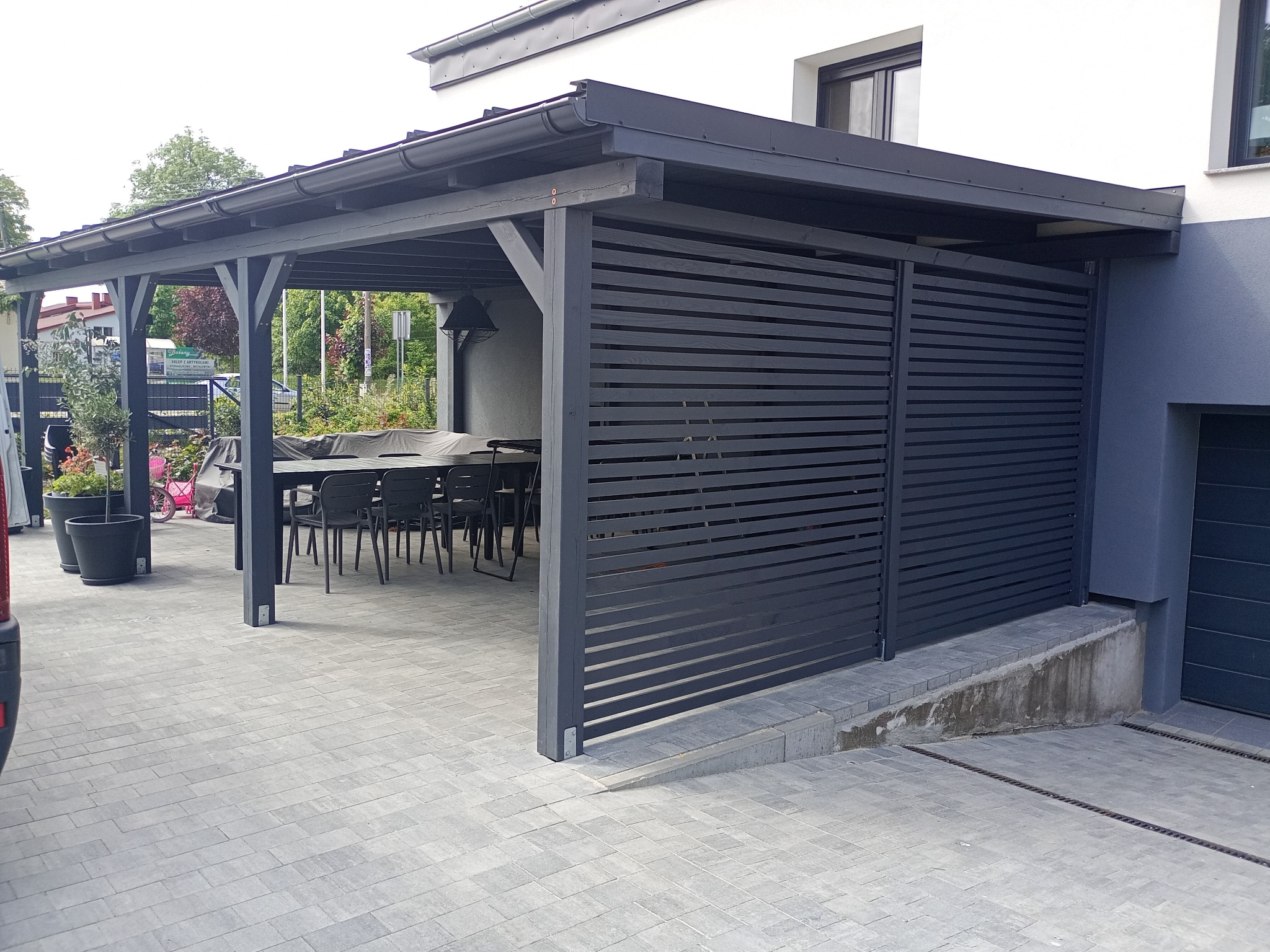 Zadaszona, szara altana z drewnianymi panelami i stołem z krzesłami na kostce brukowej. Nowoczesny design, widok z zewnątrz, częściowo widoczny samochód.