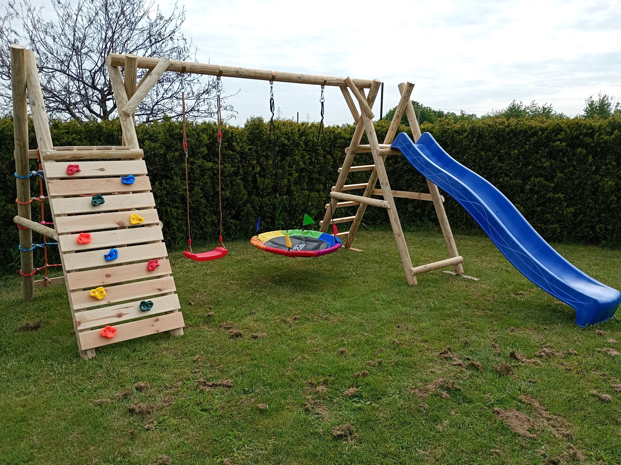 Drewniany plac zabaw z huśtawkami, ścianką wspinaczkową i zjeżdżalnią na trawniku. Tło stanowi żywopłot. Widoczne ślady po kretowiskach na trawie.