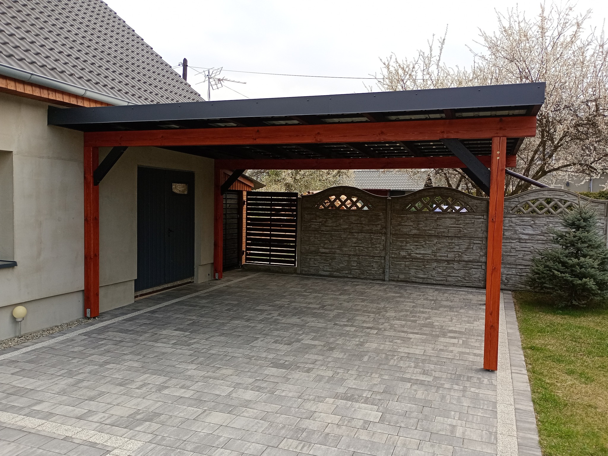 Zadaszone miejsce parkingowe z drewnianą konstrukcją i szarą kostką brukową. W tle widoczny fragment domu z szarą elewacją i ogrodzenie.