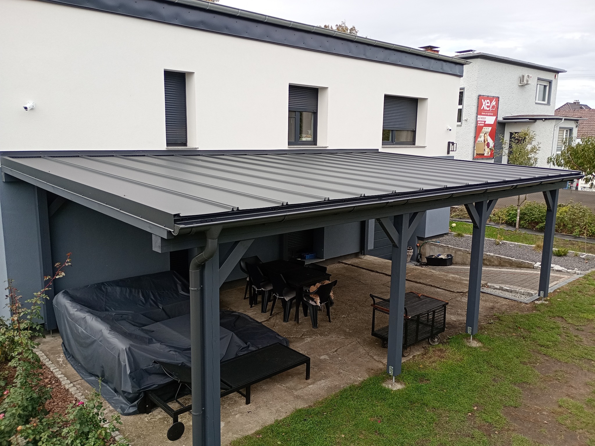 Zadaszenie tarasu w szarym kolorze, przylegające do białego budynku z roletami. Widoczne meble ogrodowe i fragment trawnika. Nowoczesna konstrukcja.