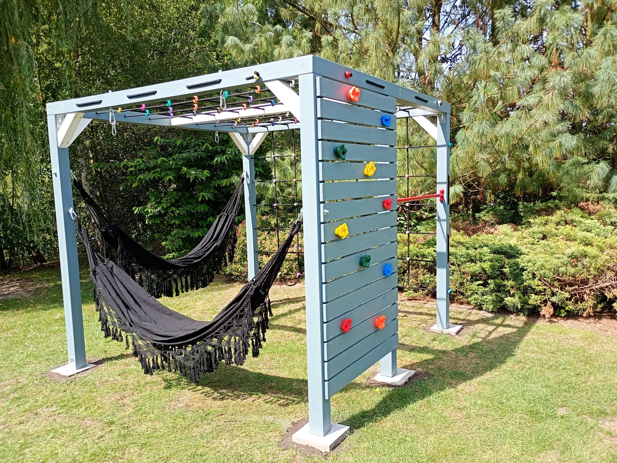 Drewniany plac zabaw z drabinkami, ścianką wspinaczkową i hamakiem na zielonym trawniku w słoneczny dzień. Elementy pomalowane na szaro-niebieski kolor.