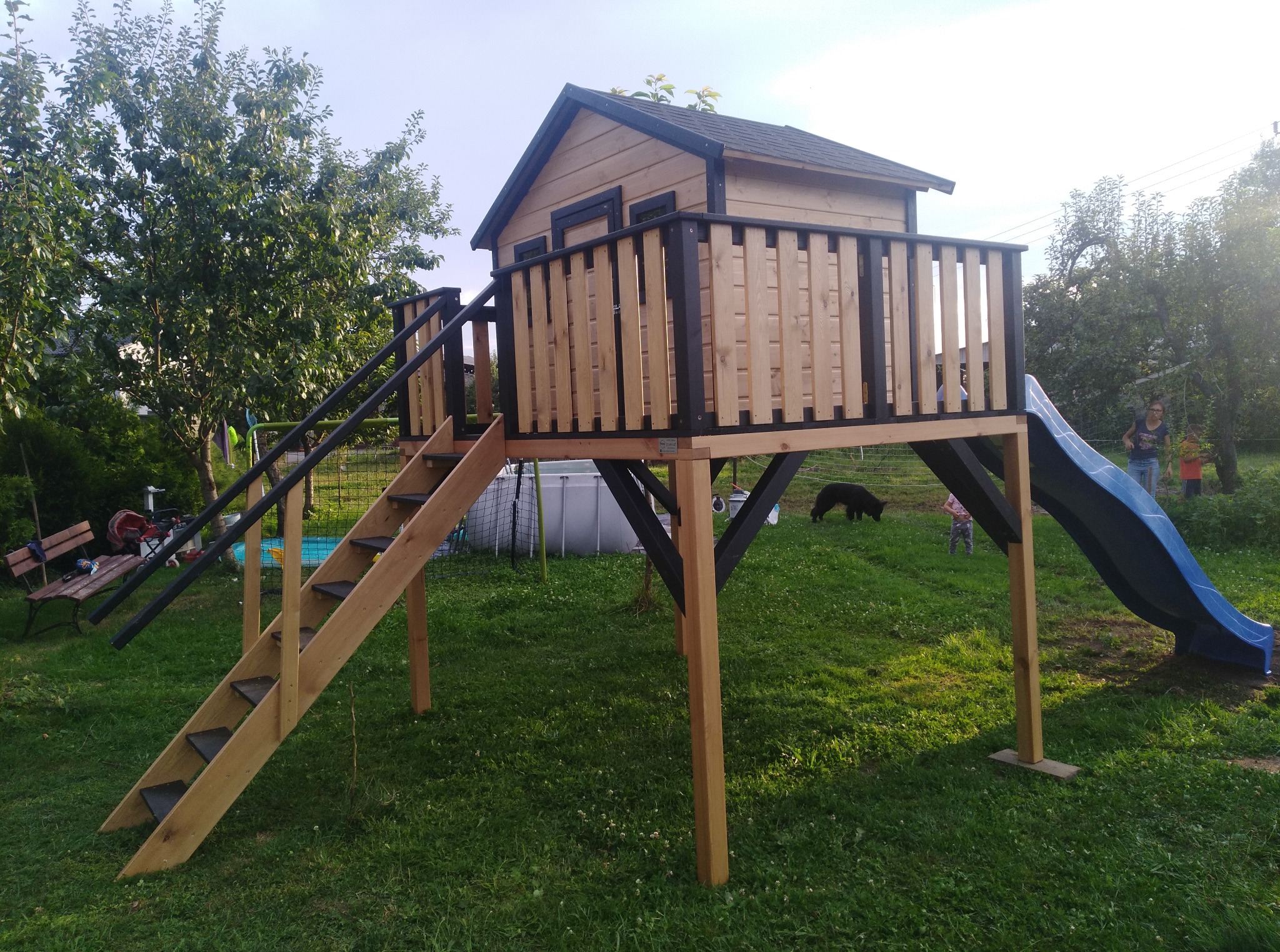 Drewniany domek na palach z czarnymi akcentami, schodami i zjeżdżalnią na trawniku. W tle drzewa i osoby, tworzące przyjazną przestrzeń do zabawy na świeżym powietrzu.