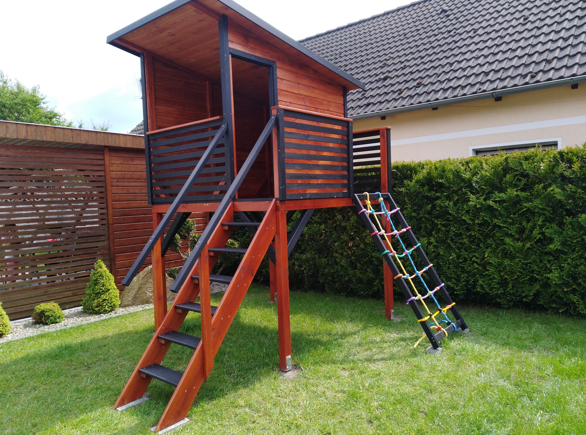 Drewniany plac zabaw z domkiem na palach, schodami i siatką do wspinaczki, umieszczony na trawniku w ogrodzie. Widoczne ogrodzenie i budynek w tle.