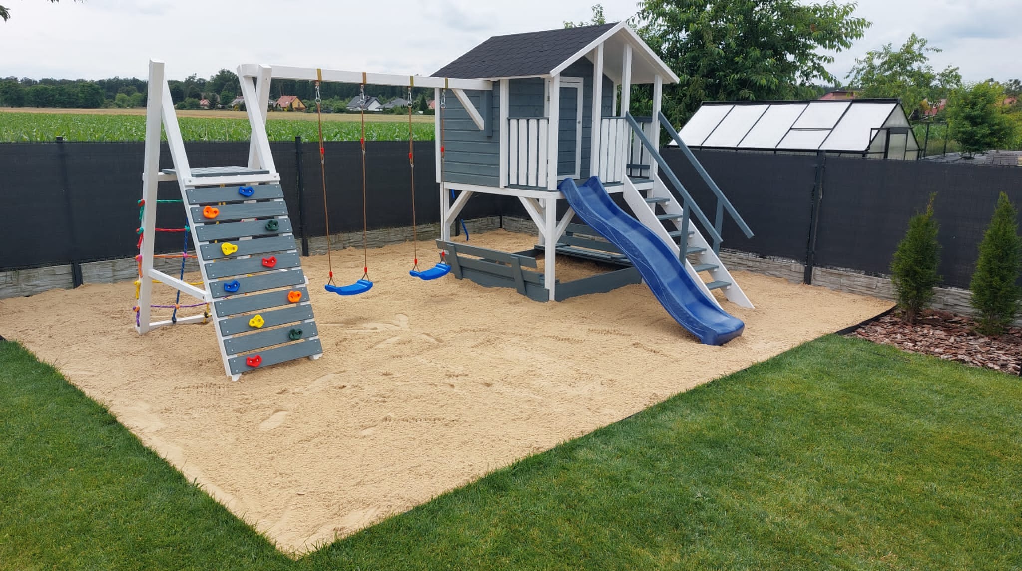 Kompleksowy plac zabaw z domkiem, zjeżdżalnią, ścianką wspinaczkową i huśtawkami, otoczony piaskiem i zielonym trawnikiem. Widok na ogród i szklarnię w tle.