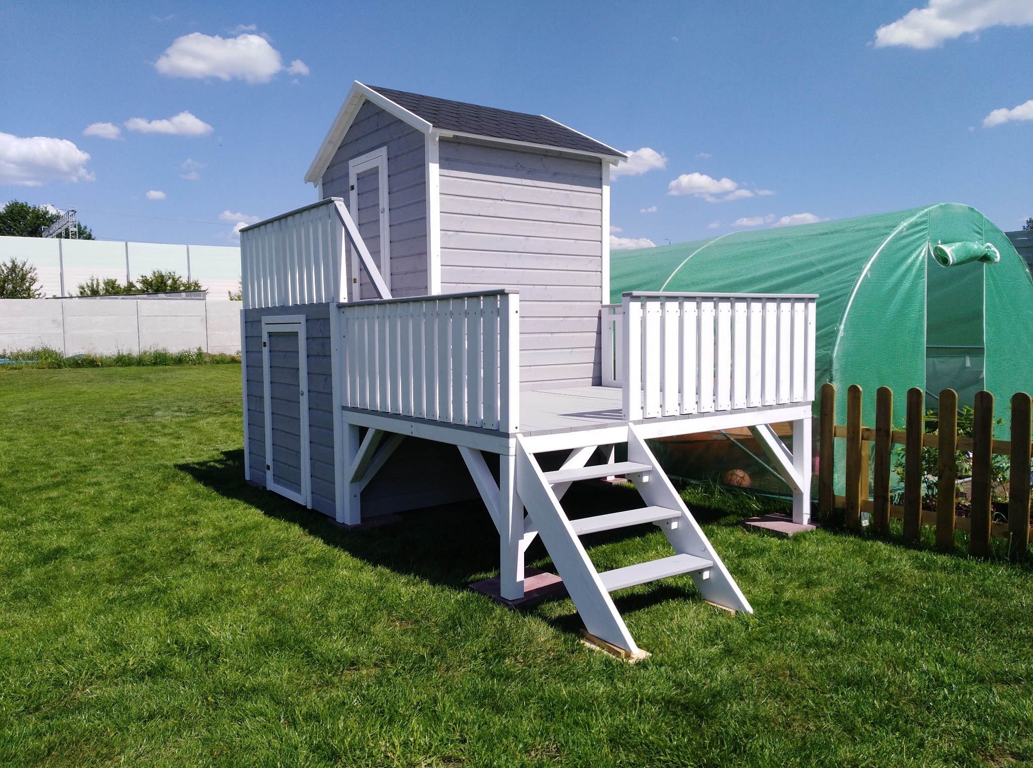 Drewniany domek zabaw dla dzieci w szaro-białej kolorystyce, z małym tarasem i schodkami, ustawiony na zielonym trawniku w słoneczny dzień. W tle szklarnia.