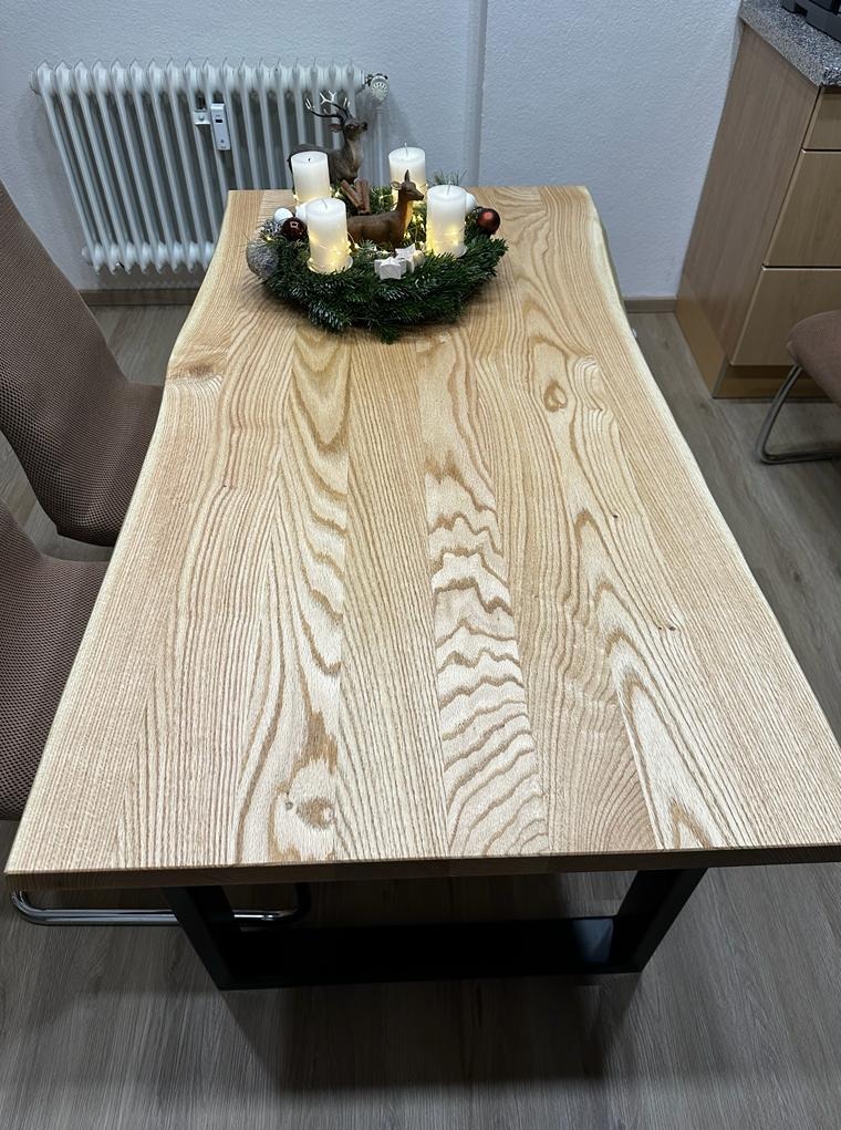 Drewniany stół z blatem o nieregularnych krawędziach, na metalowych nogach. Na stole wieniec adwentowy ze świecami. W tle grzejnik i fragment szafki.