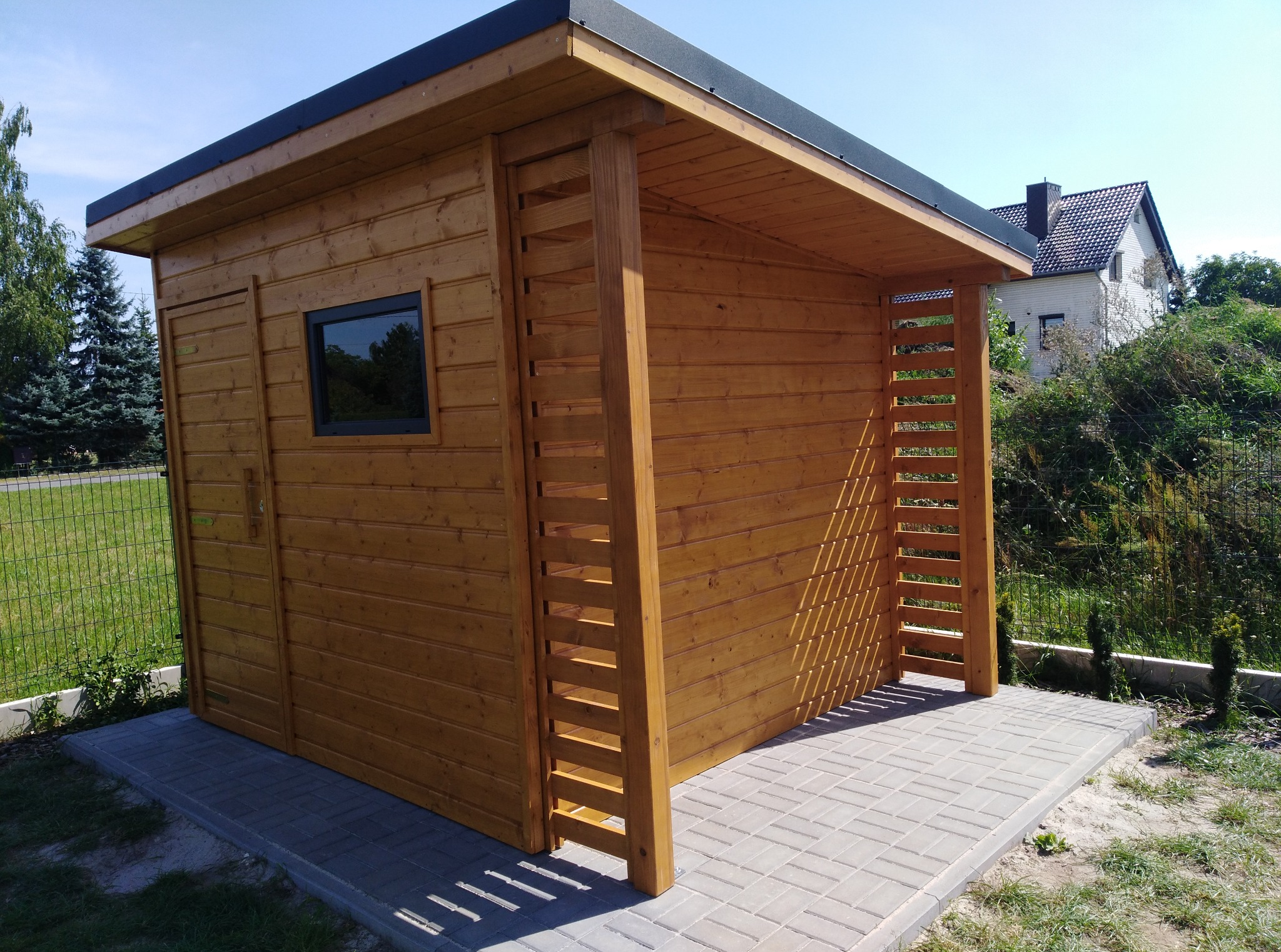 Drewniana altana ogrodowa z oknem, na wybrukowanym podeście, w tle zieleń i dom. Konstrukcja z belek i desek, zadaszona, w naturalnym odcieniu drewna.