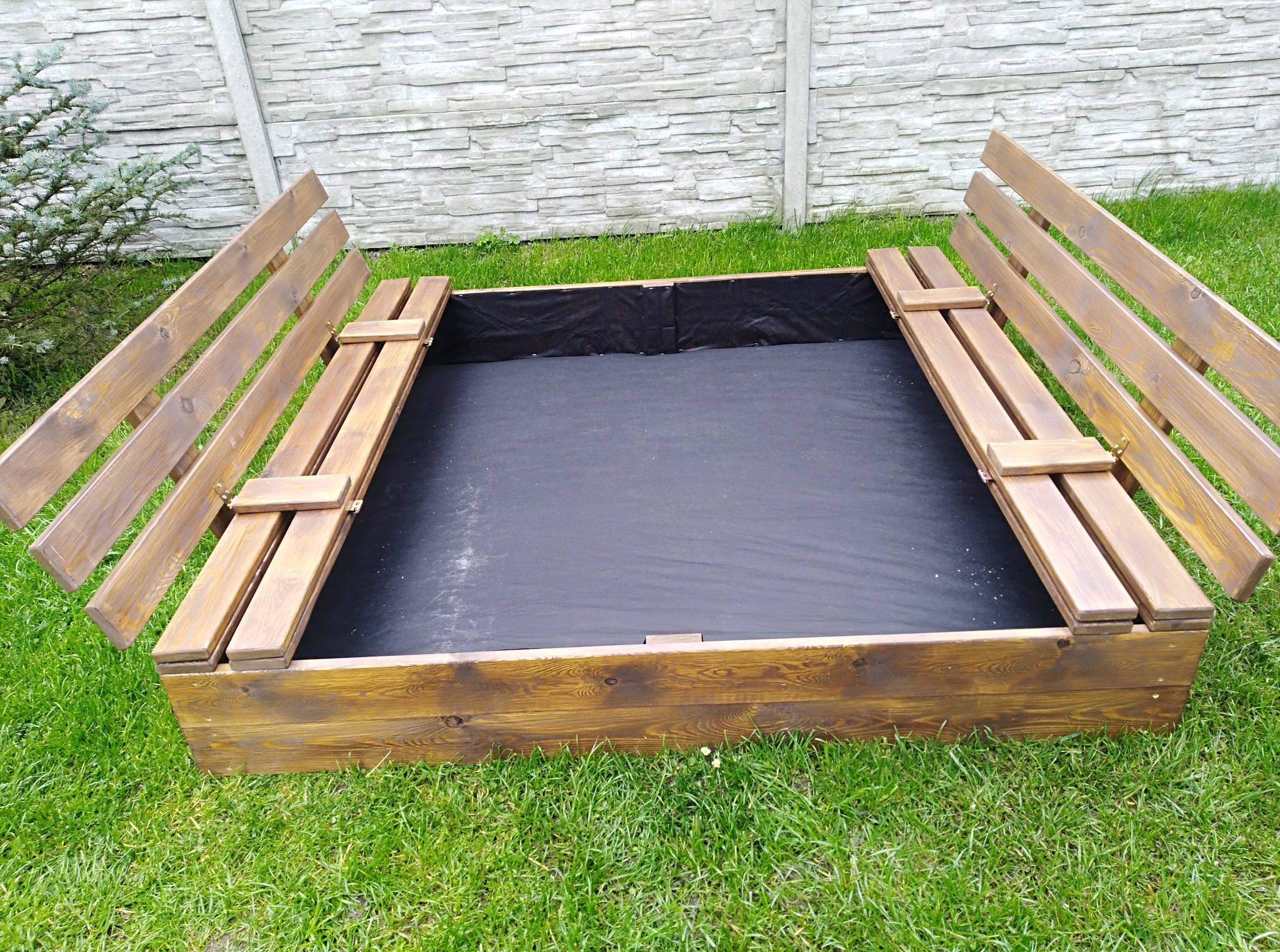 Drewniana piaskownica z rozkładanymi ławeczkami na trawie, wyłożona czarną tkaniną. Widoczny fragment ogrodzenia z betonu architektonicznego i iglak.