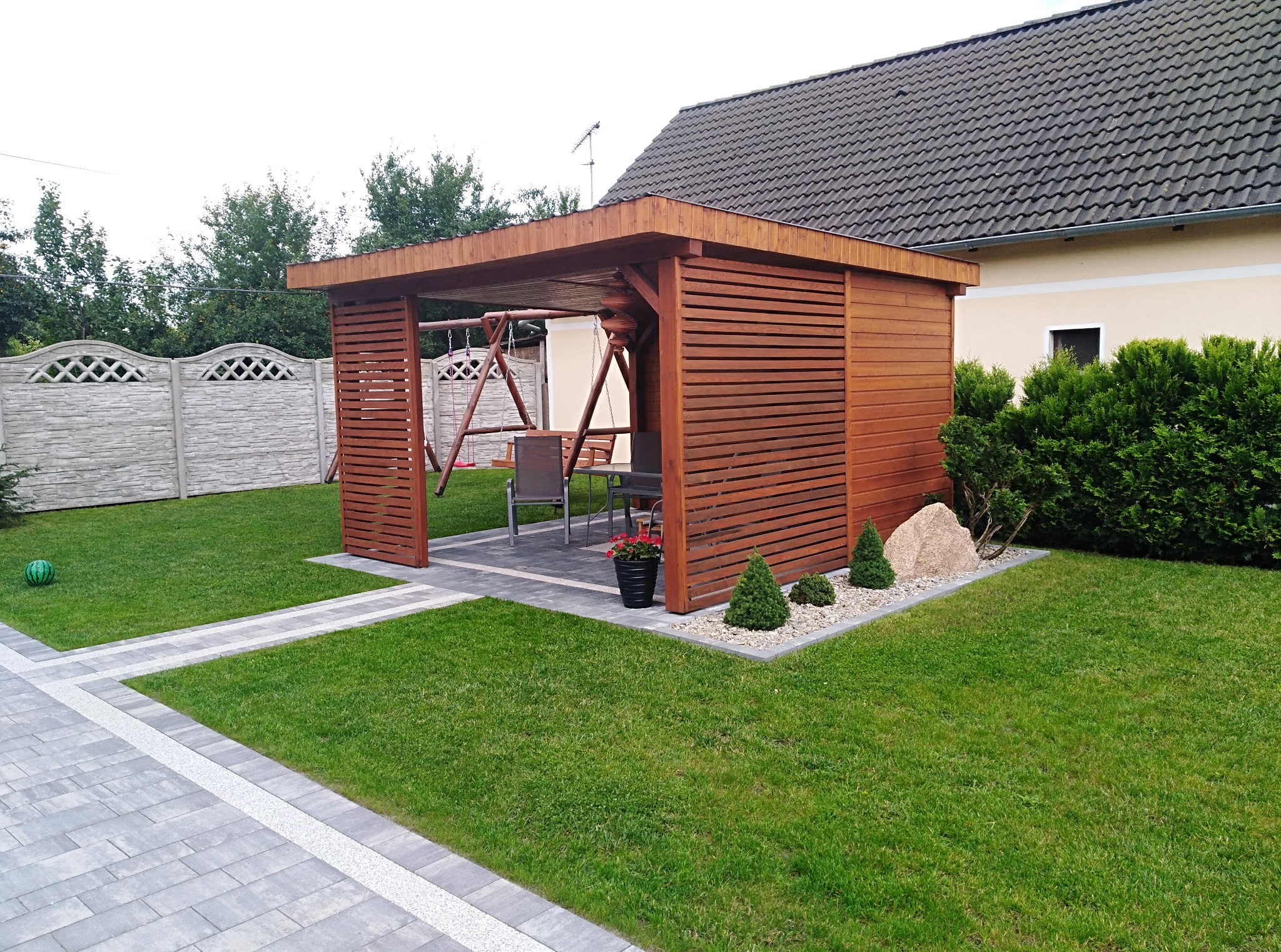 Drewniana altana z huśtawką i meblami ogrodowymi na trawniku w Starych Siołkowicach. Obok ścieżka z kostki brukowej i zadbana zieleń.