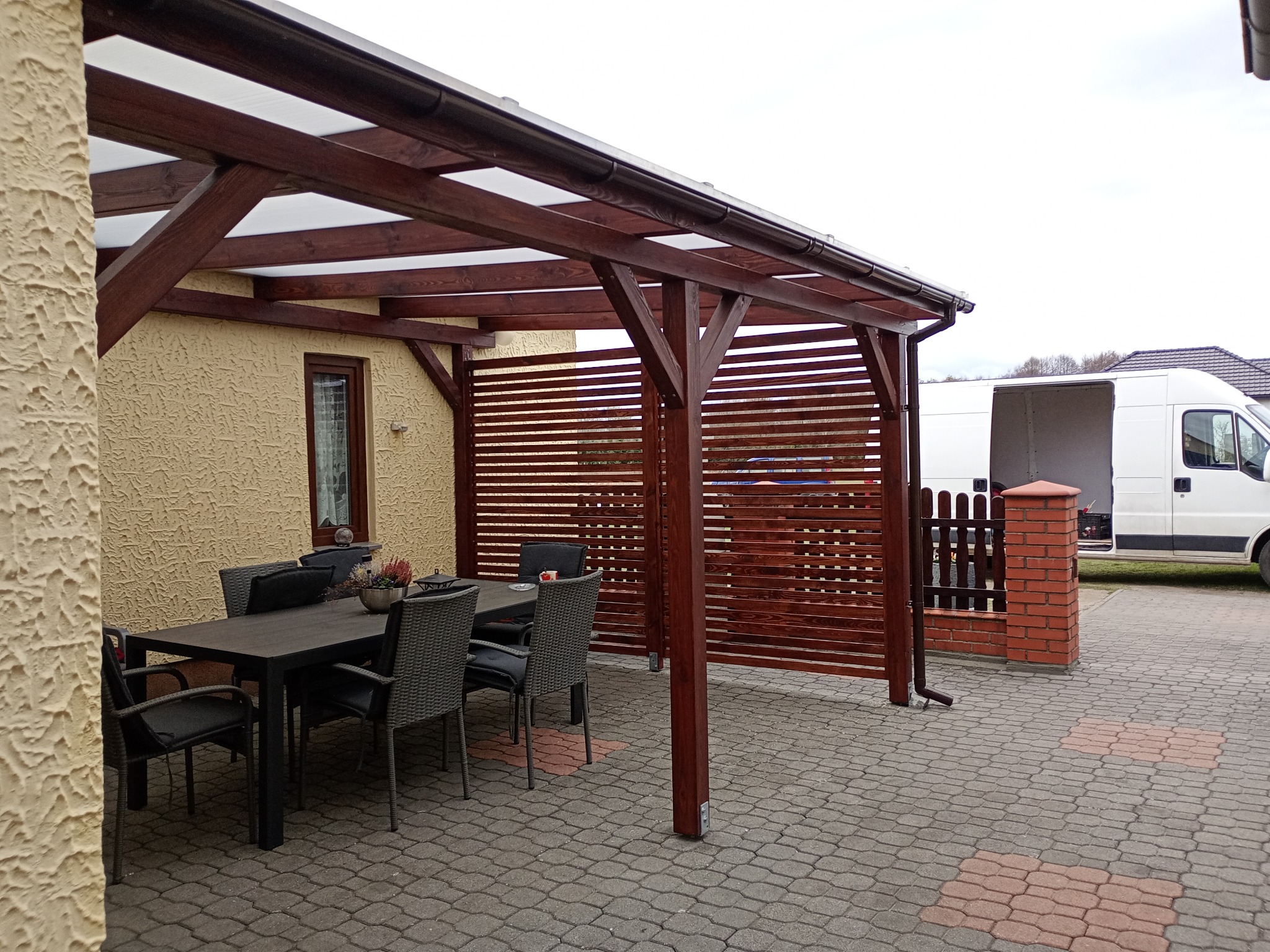 Zadaszona drewniana pergola z meblami ogrodowymi na brukowanym patio. W tle biały bus dostawczy z otwartymi drzwiami. Widok na zewnątrz, pochmurne niebo.